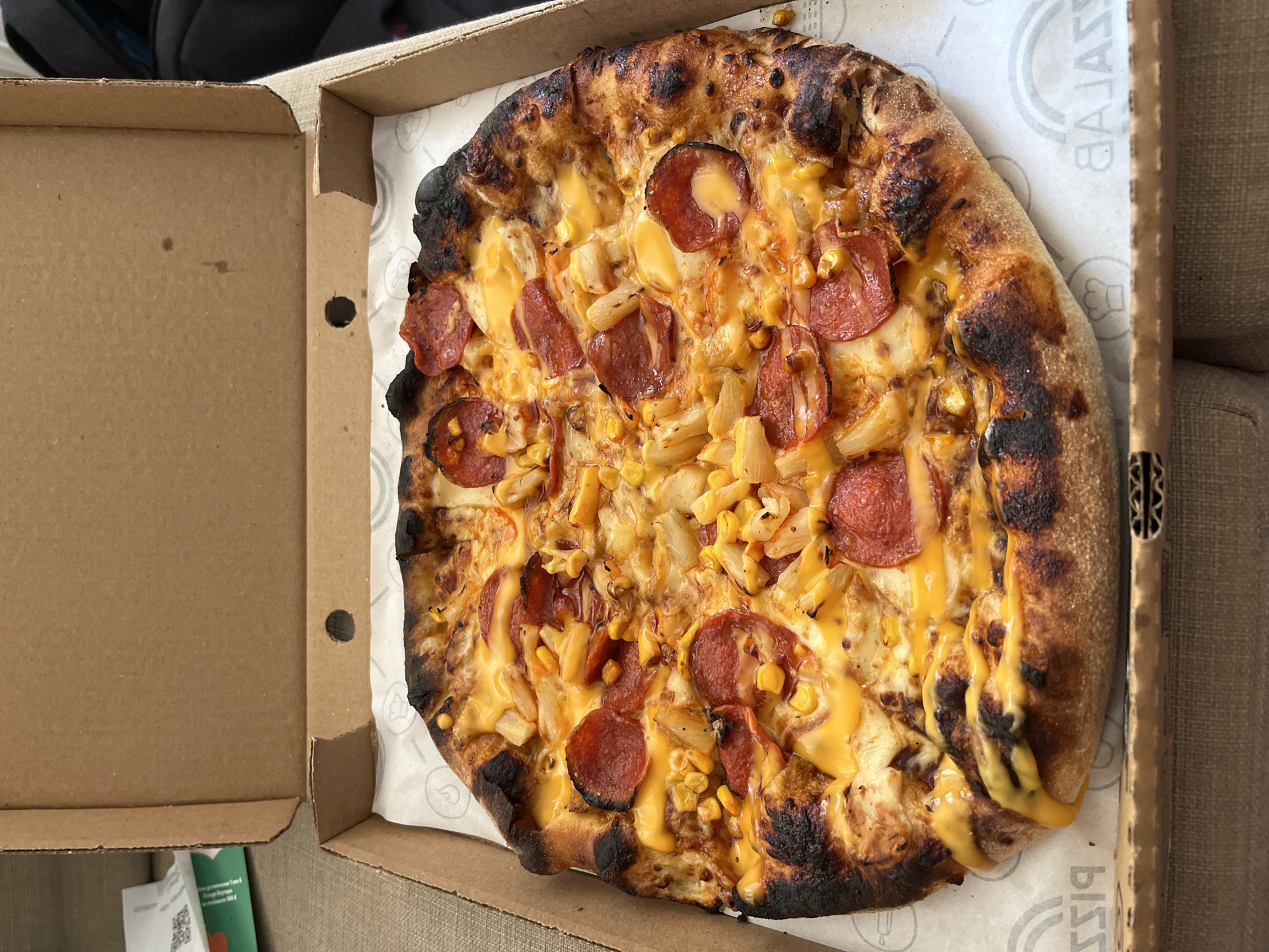 A close-up, overhead shot shows a pepperoni and corn pizza in a cardboard pizza box. The pizza has a slightly charred crust and is topped with melted cheese, pepperoni slices, corn kernels, and what appears to be pineapple. The pizza is placed on a piece of white paper with a repeating circular logo. The pizza box is open, revealing the pizza and the white paper liner. The box itself is made of brown cardboard. In the background, to the left, is the inside of the pizza box lid, and to the right, part of a couch or chair is visible. The lighting suggests daytime. The setting appears to be indoors, possibly a home or a casual dining area. The pizza looks freshly prepared, suggesting it might have been delivered or just taken out of the oven. There is visible text on the white paper liner, including the word "GALAXY" in a circular logo. There is also some text visible on a small green flyer at the bottom left corner of the image, which is blurry but appears to be in Bulgarian.