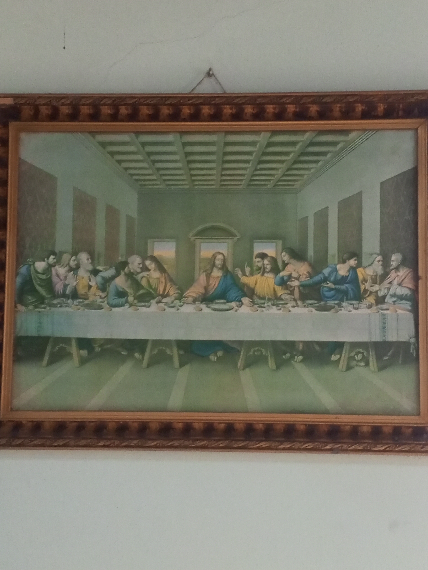 This is a framed painting of "The Last Supper." The scene depicts Jesus Christ at the center of a long table, surrounded by his twelve apostles. They are gathered for a meal, with Jesus gesturing with his right hand. The figures are dressed in historical attire, and the setting appears to be an interior room with a patterned ceiling and arched doorways in the background. The painting is rendered in a classical style. The frame is ornate and golden-brown. The painting is hung on a white wall, with visible wall imperfections and a hanging wire at the top. The lighting suggests daytime.