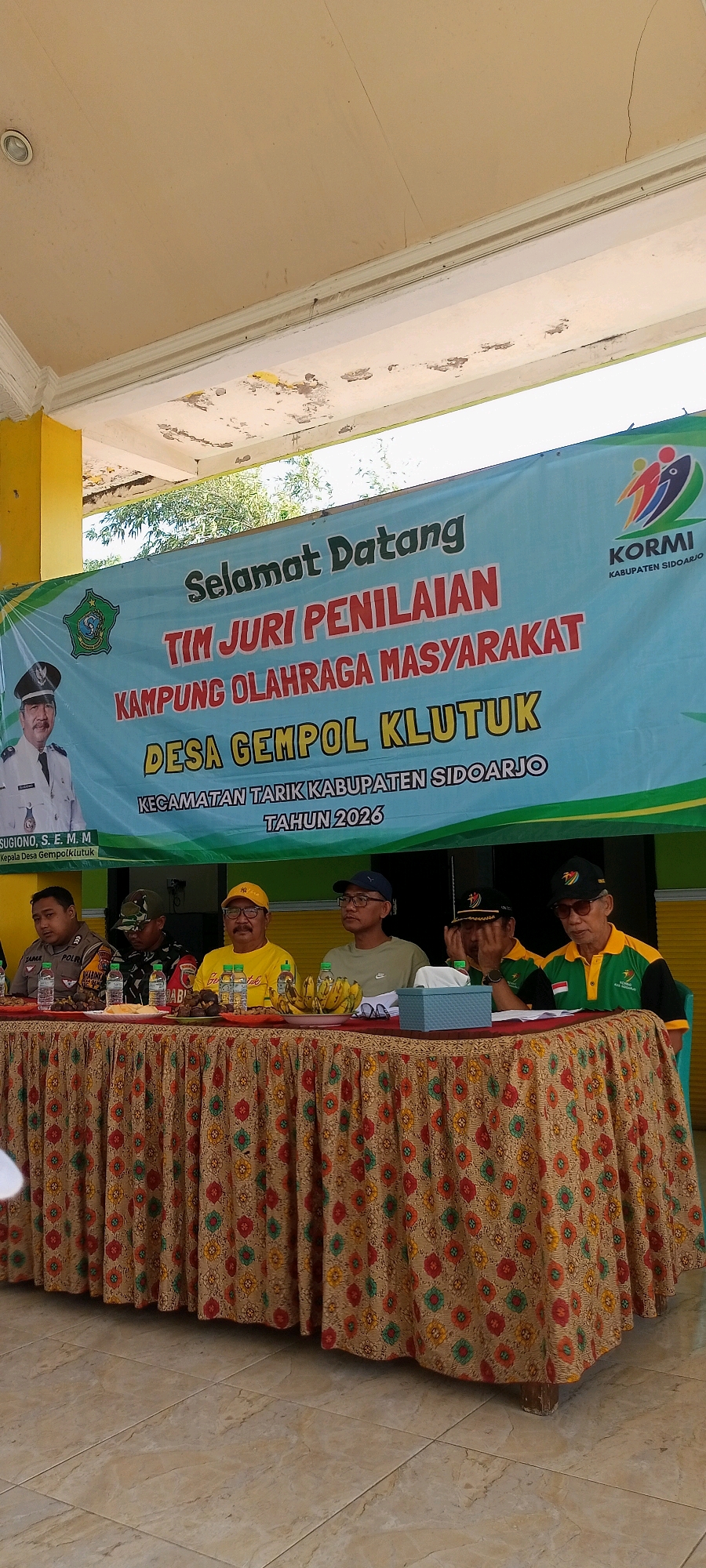 A group of men are seated behind a table covered with a patterned cloth. The table displays food items and drinks. In the background, a banner reads "SELAMAT DATANG TIM JURI PENILAIAN KAMPUNG OLAHRAGA MASYARAKAT DESA GEMPOL KLUTUK KECAMATAN TARIK KABUPATEN SIDOARJO TAHUN 2026". To the left of the banner, there is a portrait of a man in uniform. The setting appears to be outdoors, under a canopy.