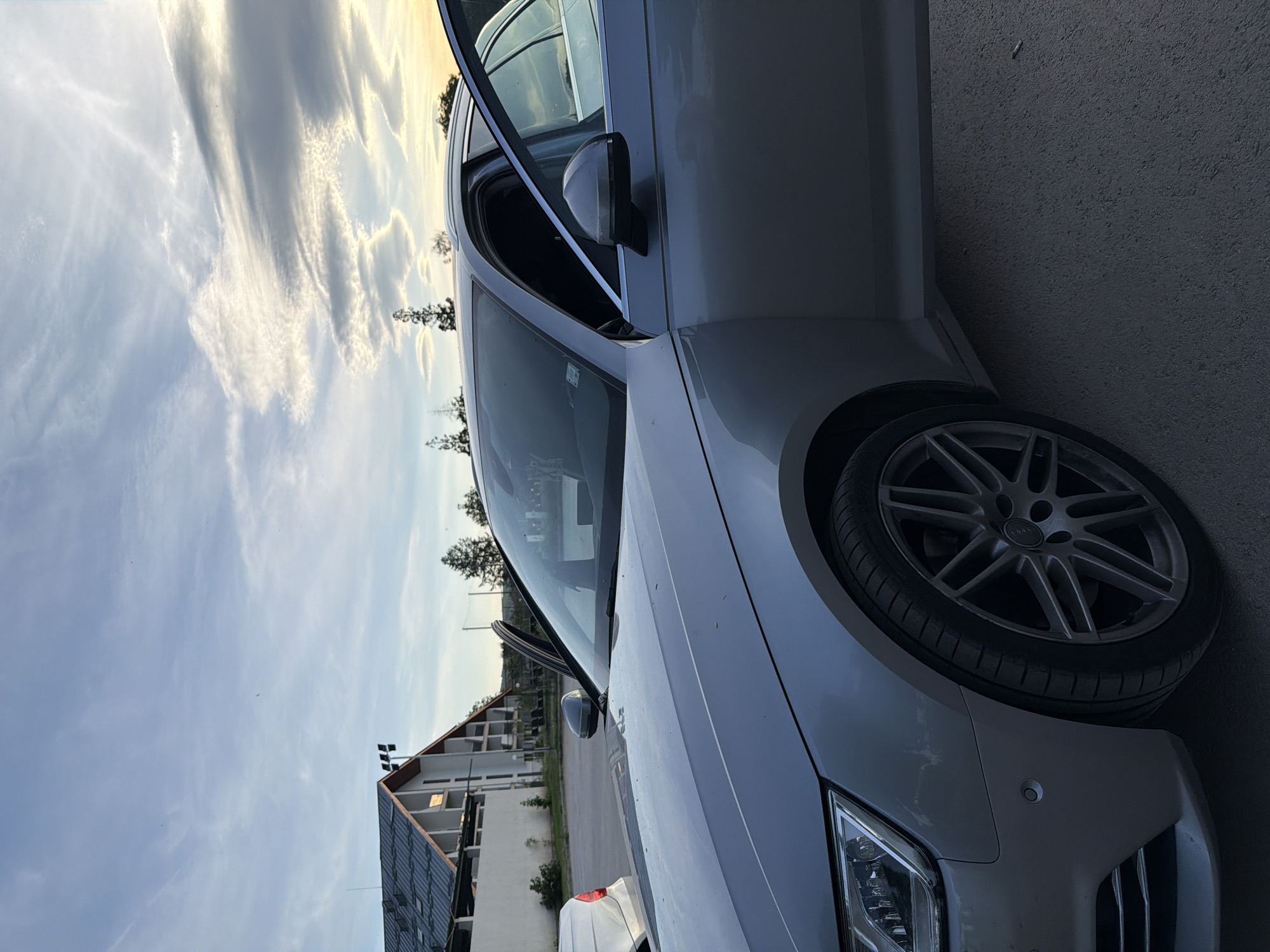 A white Audi car is parked on an asphalt surface. The front passenger door is open. In the background is a building with solar panels, indicating it is likely a modern or renovated structure. The sky is visible with clouds. The location is near a building and a road, indicating an urban or suburban setting, potentially in Panagyurishte, Bulgaria. The image does not include people or any specific actions.
