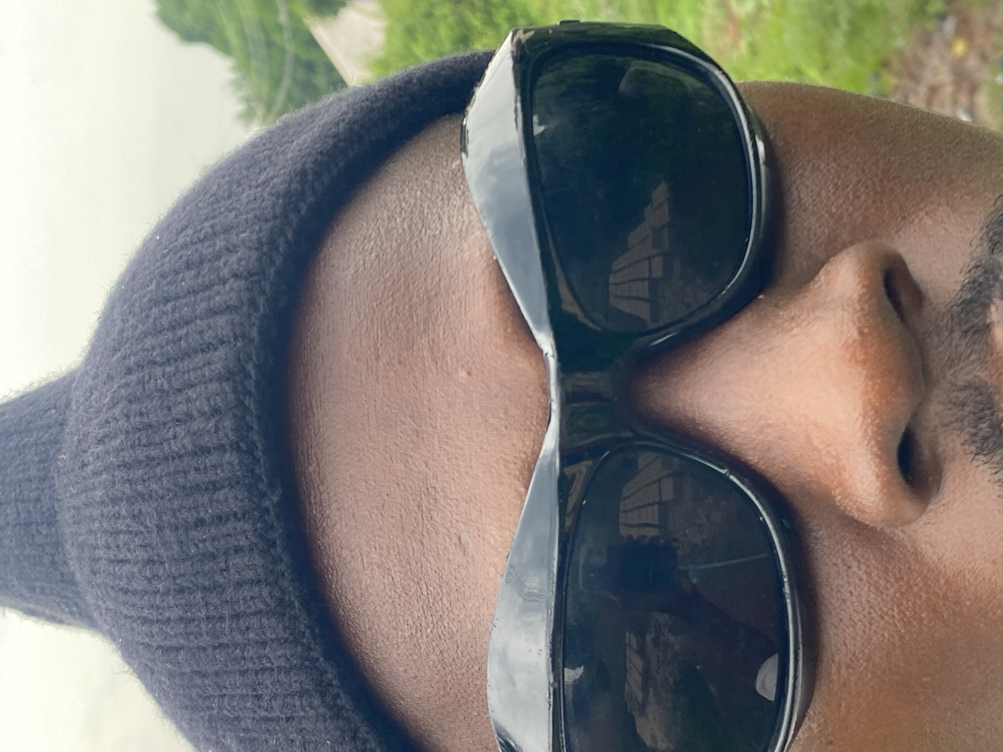 A close-up, rotated view of a person wearing a dark beanie and sunglasses. The person has dark skin and a beard, and their nose is prominently featured in the frame. The sunglasses reflect a scene outside, possibly a building and greenery. The background suggests an outdoor setting, though the exact location in Amaigbo, Nigeria is not discernible from this image.