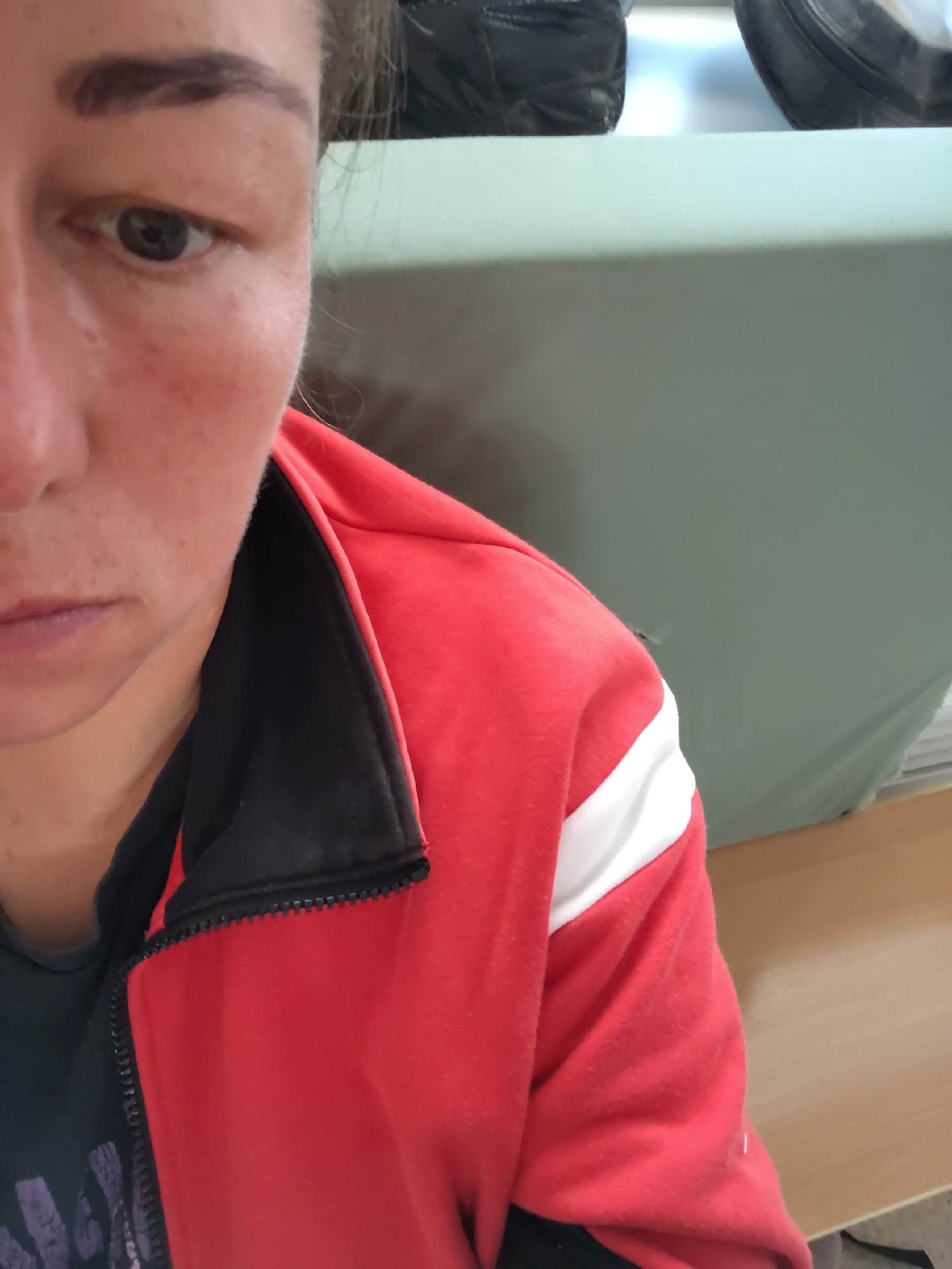 A close-up shot shows the left side of a person's face and upper body. The person has fair skin and dark eyebrows. They are wearing a red zip-up jacket with black trim and a white stripe across the chest. The background is blurred and consists of a light green surface and some dark objects. There is no information about the location of Chaikovsky, Russia.
