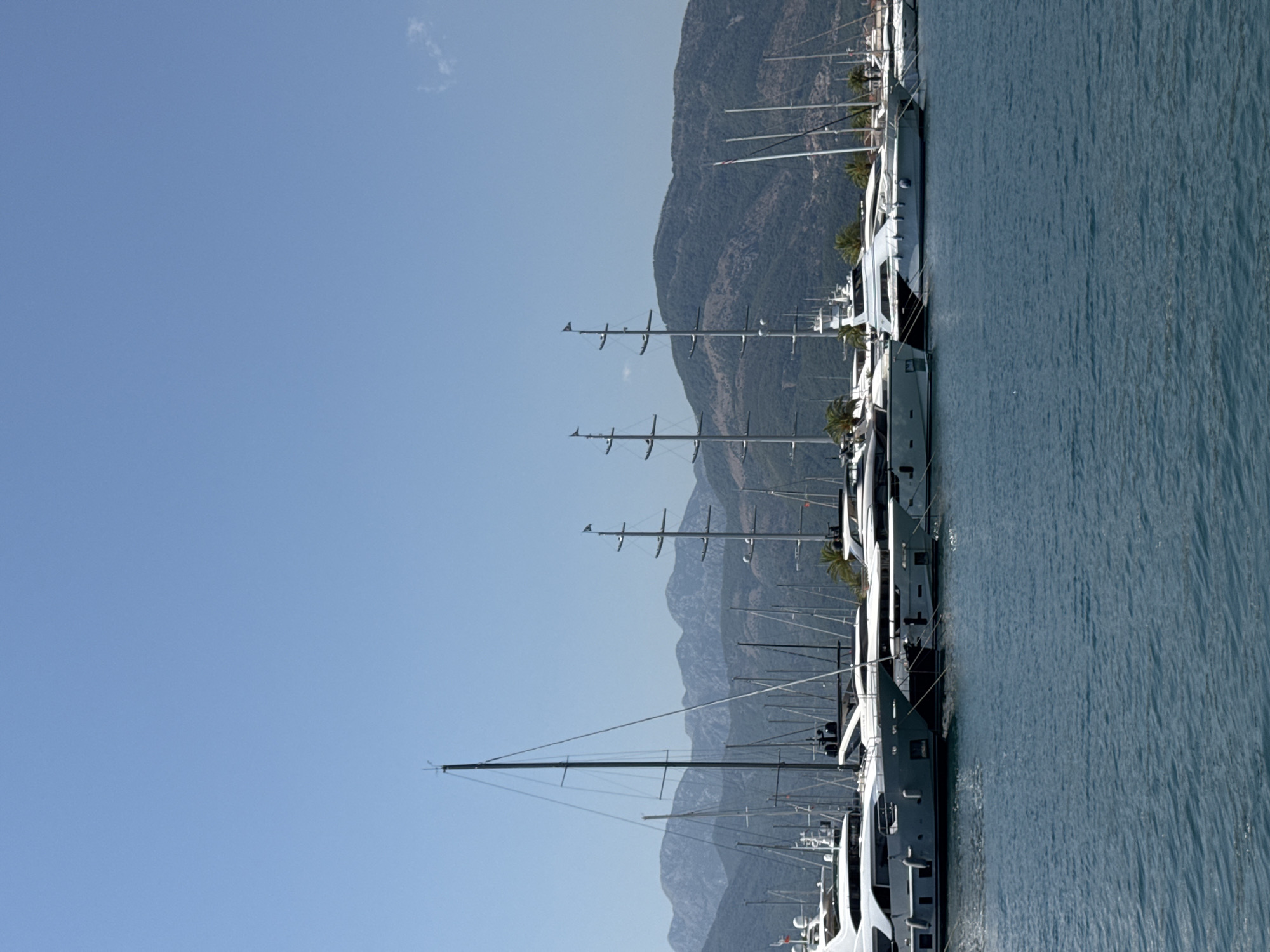 In Tivat, Montenegro, a marina is depicted on a clear day under a bright blue sky. The foreground is dominated by the blue water of the bay, with gentle ripples on the surface. Moored in the middle ground are numerous yachts of varying sizes, mostly white and gray, lined up next to each other. Some of these are large motor yachts with sleek designs, while others appear to be sailboats with tall masts. In the background, lush green mountains rise, their slopes covered in dense vegetation, and the peaks are textured with rocky outcrops. The scene captures a serene maritime setting.