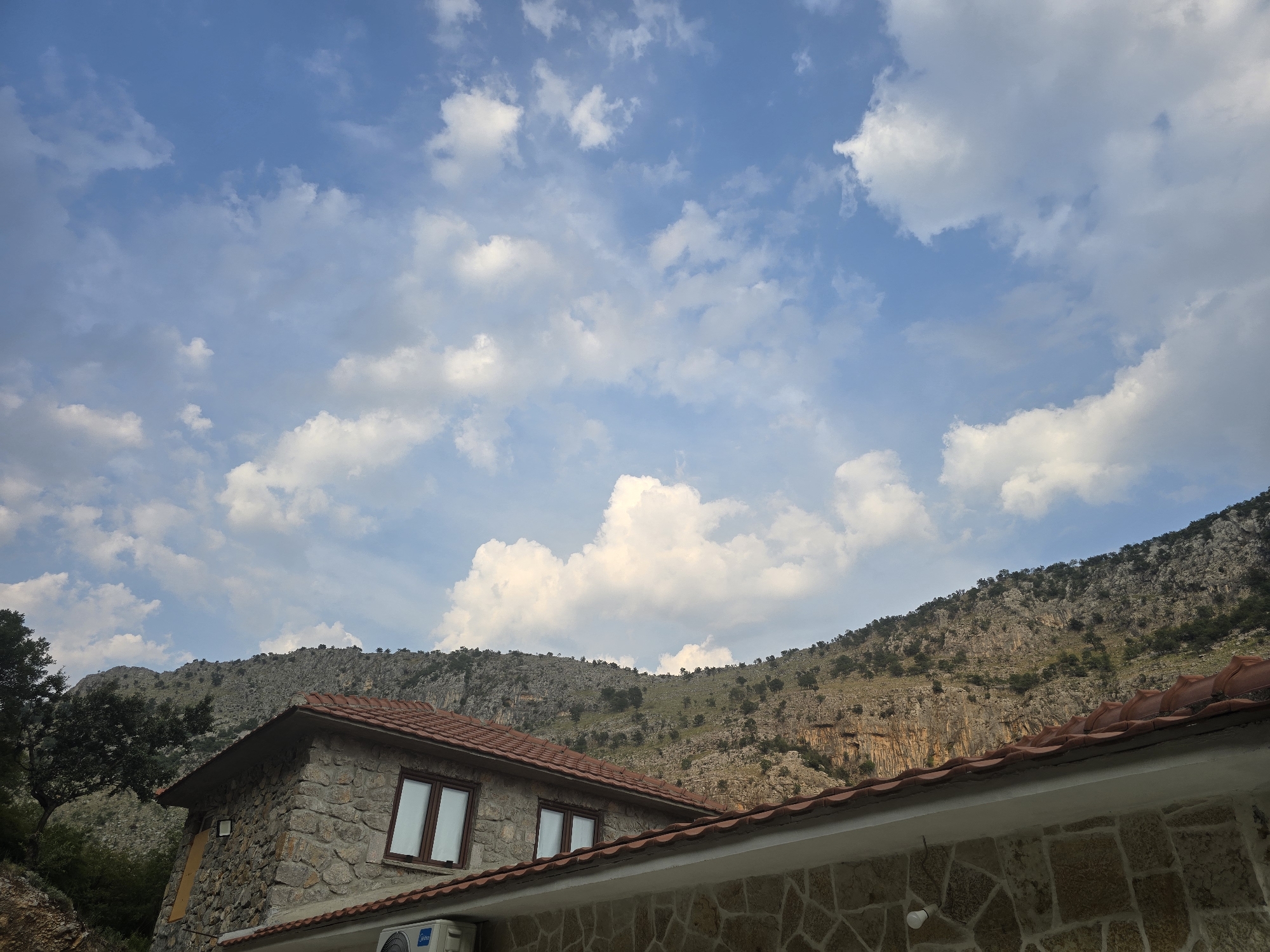 The image depicts a stone house with a tiled roof, nestled against a rocky, mountainous backdrop under a partly cloudy blue sky. In the foreground, a portion of the house's exterior is visible, constructed from rough-hewn stones. Two windows with dark frames are present on the visible wall, and an air conditioning unit is mounted below one of them. The roof has a reddish-brown tile covering, with the edge of another structure visible in the lower right corner. The middle ground is dominated by the imposing mountain range, characterized by its rugged, stony surface dotted with sparse vegetation. A large, leafy tree stands to the left of the house, adding a touch of green to the scene. The sky is a vibrant blue, scattered with fluffy white clouds, suggesting fair weather. The overall impression is one of a secluded, rustic dwelling in a mountainous region. Without specific landmarks or signage, the exact location within Podgorica, Montenegro, cannot be definitively determined from the image alone, though the terrain suggests an area on the outskirts of the city, possibly in a more rural or natural setting.