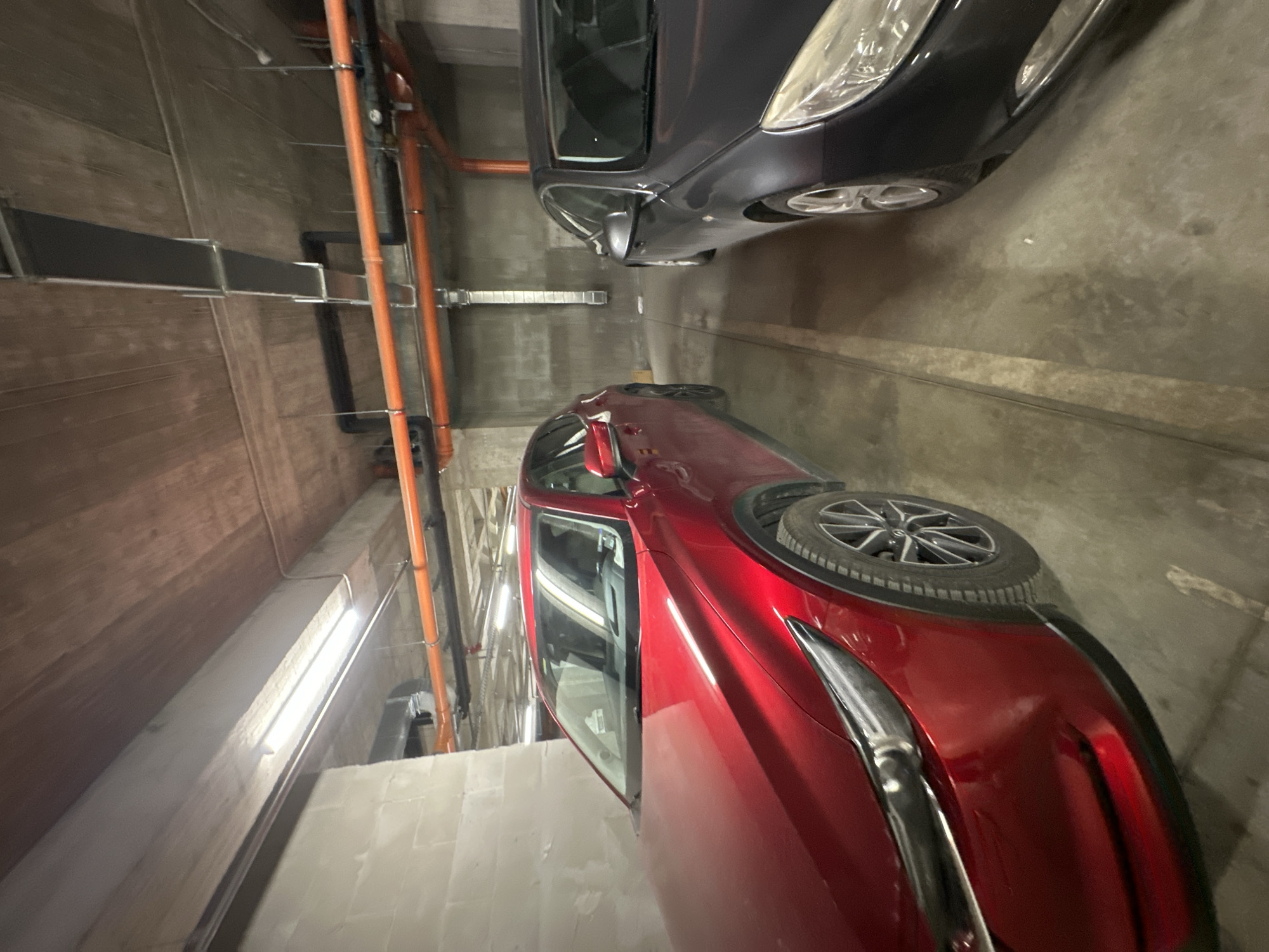 The image depicts a parking garage, likely an indoor or underground structure, with two cars parked within. One car is dark in color (possibly black or a dark grey) and appears to be parked normally. The second car, which is red, is prominently featured and angled towards the viewer; it also has a spare wheel strapped to the back of the car. Utility pipes run along the side walls of the garage. The location is likely a parking garage but without specific information within the frame the city of Varna, Bulgaria cannot be determined.