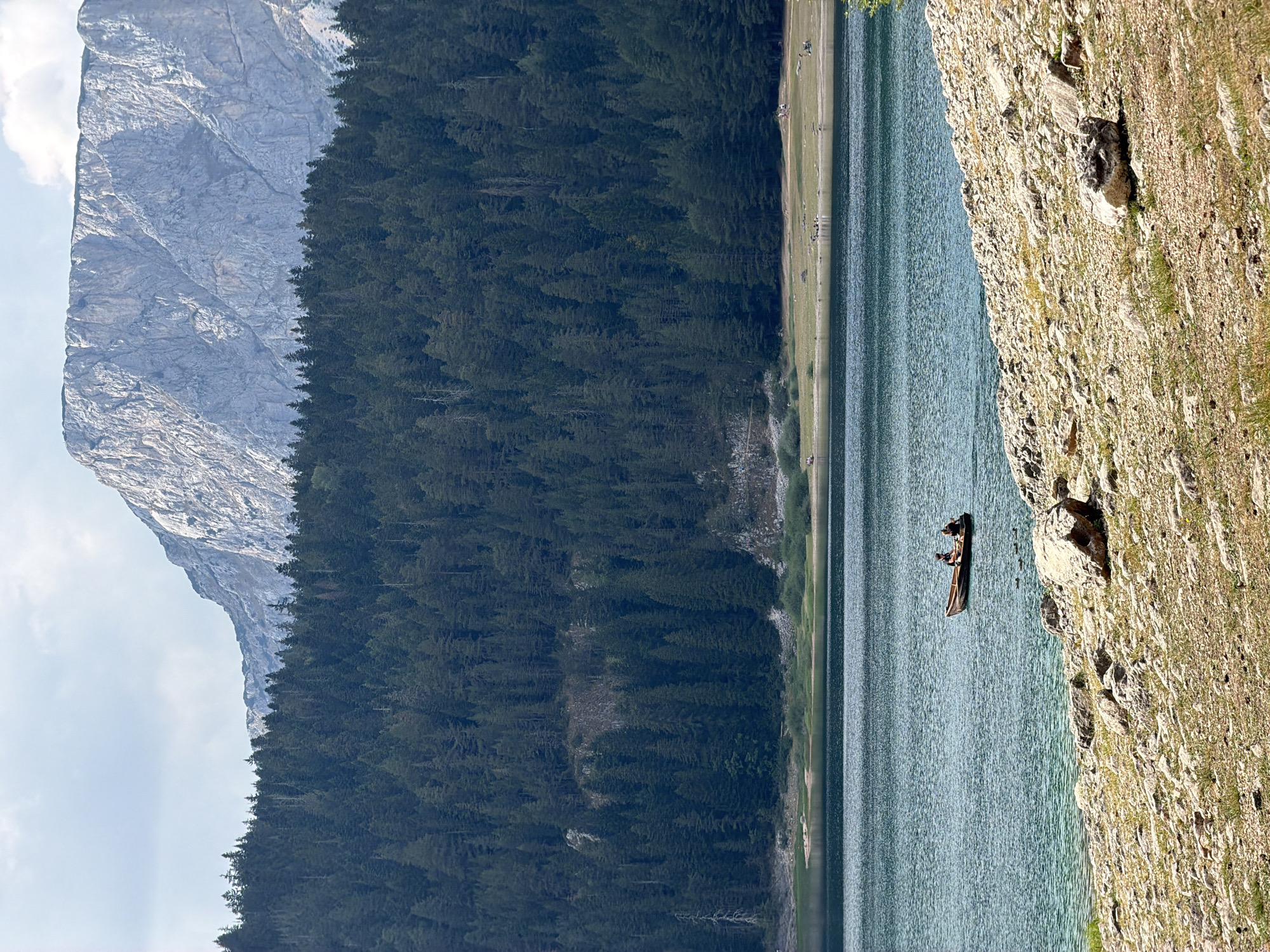 The image depicts a scenic view of Black Lake (Crno Jezero) in the Durmitor National Park, near the village of Pitomine, Montenegro. In the foreground, a rocky shoreline leads to the clear, blue-green waters of the lake. Two individuals are visible in a small wooden boat on the lake, appearing to be fishing or enjoying a leisurely row. Further in the background, a dense, dark green forest covers the lower slopes of a majestic, rugged mountain. The mountain's imposing grey rock face, etched with shadows, dominates the upper portion of the image, with a bright, cloudy sky above. The overall scene conveys a sense of natural beauty and tranquility.