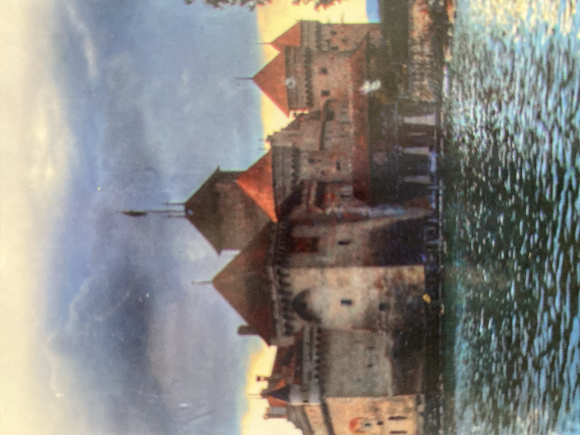 A painting depicts a stone castle with red-tiled roofs situated on the edge of a body of water, possibly a lake or river. The castle features multiple towers, turrets, and crenellated battlements, indicating a historical or defensive structure. The architecture suggests a European style. The sky above is dramatic, with heavy, dark clouds interspersed with lighter, illuminated areas, creating a sense of impending weather or a moody atmosphere. The water in the foreground is choppy with reflections of the sky and the castle. There are no visible people or animals in the painting. The overall impression is one of grandeur and historical significance, set against a dramatic natural backdrop.