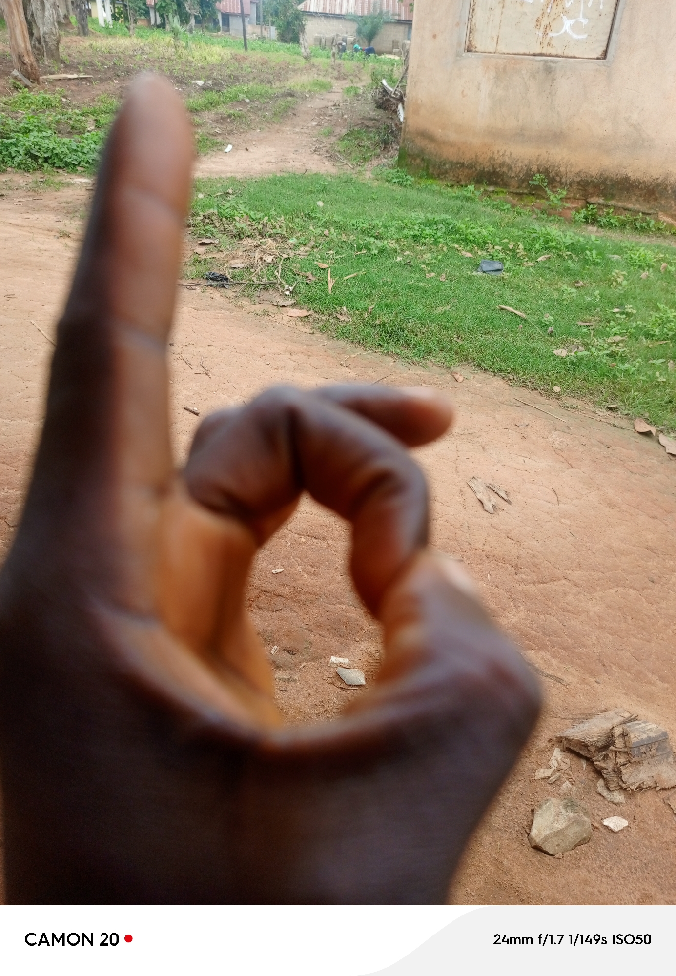 The image shows a close-up of a person's hand forming an "okay" gesture, with their thumb and index finger touching. The hand is in the foreground and is out of focus. In the background, there is a dirt path leading towards some buildings with corrugated metal roofs, suggesting a rural or semi-urban setting. Green grass grows along the sides of the path and around the buildings. A light-colored, somewhat weathered wall of a building is visible on the right side of the image. The ground appears to be dry and reddish-brown, with scattered stones and some debris. The overall scene suggests an outdoor environment during daylight. The visible text at the bottom of the image indicates "CAMON 20" and "24mm f/1.7 1/149s ISO50", which are camera model and settings information. The location context is Takau, Nigeria.