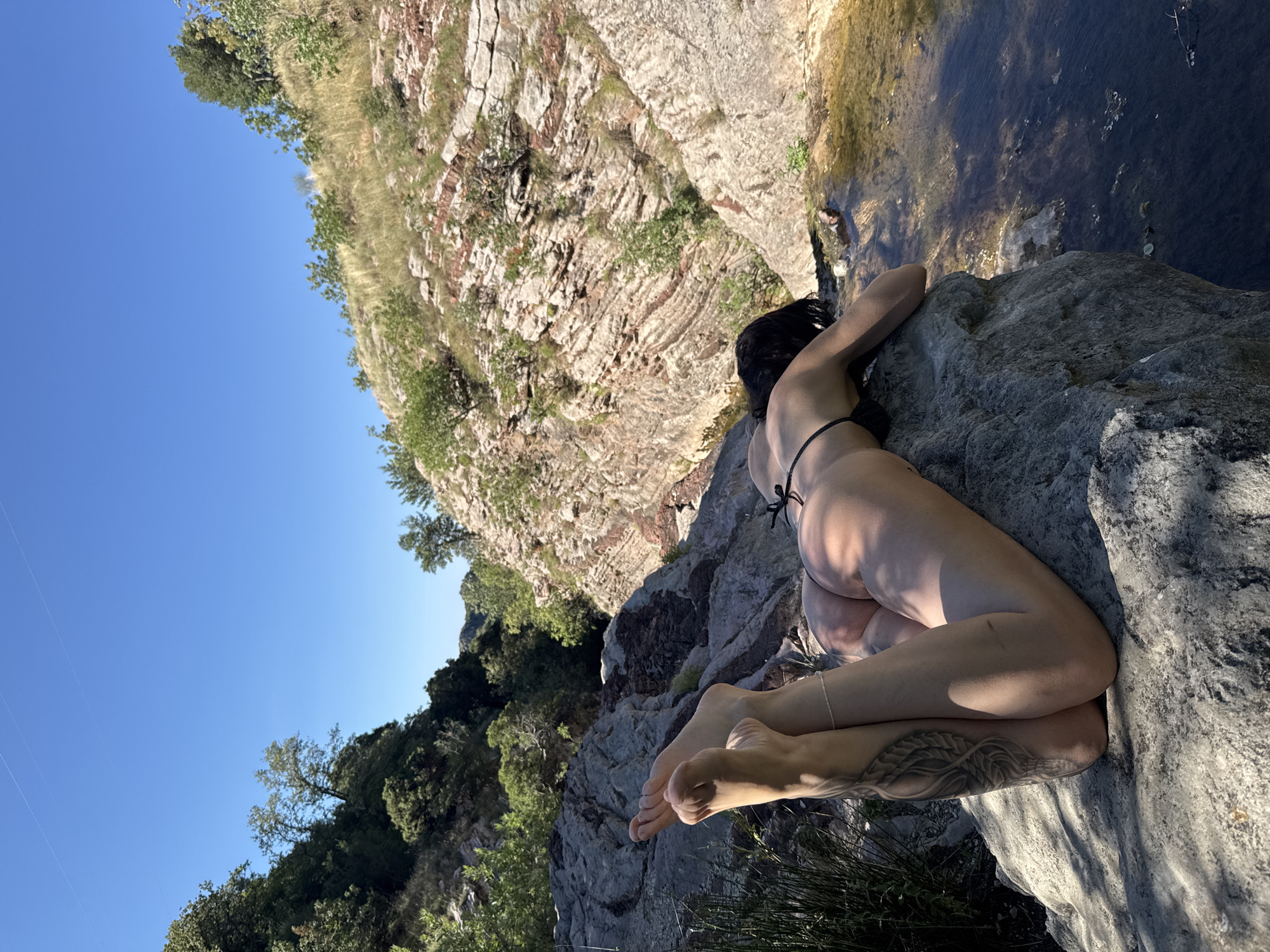 A person with dark hair lies face down on a large, grey rock, their upper body partially obscured. They are wearing a black bikini. Their legs are extended upwards, and their feet are visible, with a tattoo on the left calf. The rock is situated near a body of water with a rocky, uneven shoreline. Behind the rock, steep, layered rock formations rise up, covered in sparse vegetation and trees. The sky above is clear and blue. The scene appears to be a natural outdoor environment.