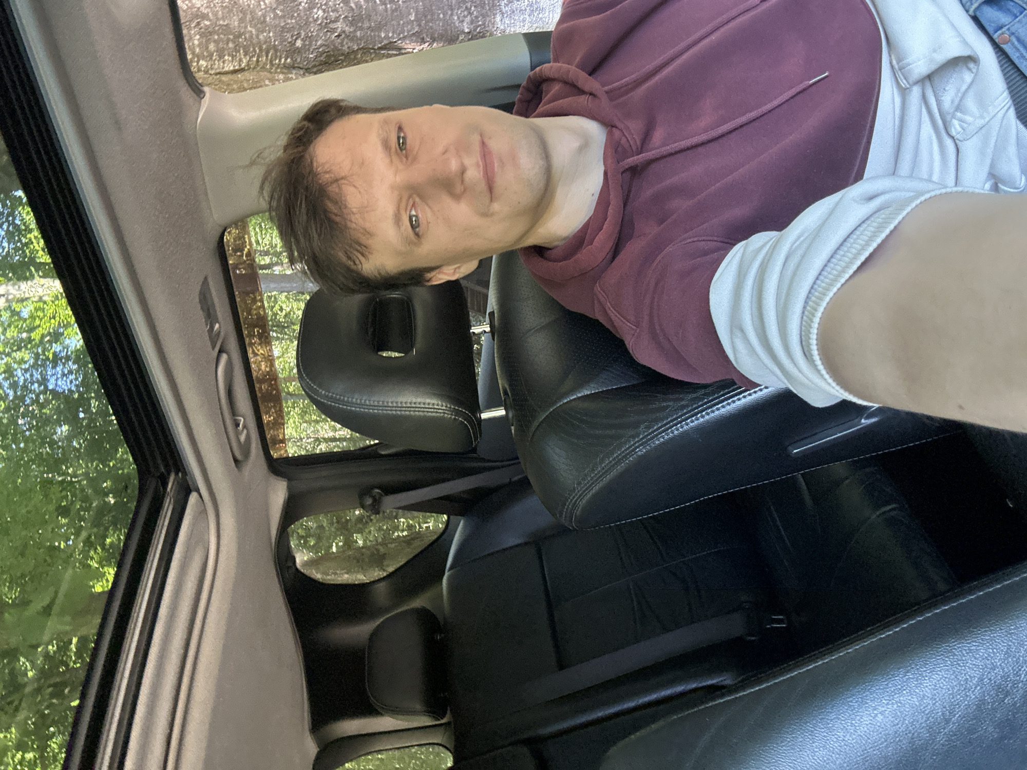 A man is seated in the front passenger seat of a car. He is wearing a maroon hooded sweatshirt and a white long-sleeved shirt underneath. The car interior is visible, featuring black leather seats and a sunroof. Through the sunroof and windows, lush green trees and foliage suggest an outdoor, possibly wooded, environment. The car is parked, and the man is looking towards the camera, taking a selfie. The location is identified as Shatava, Ukraine.
