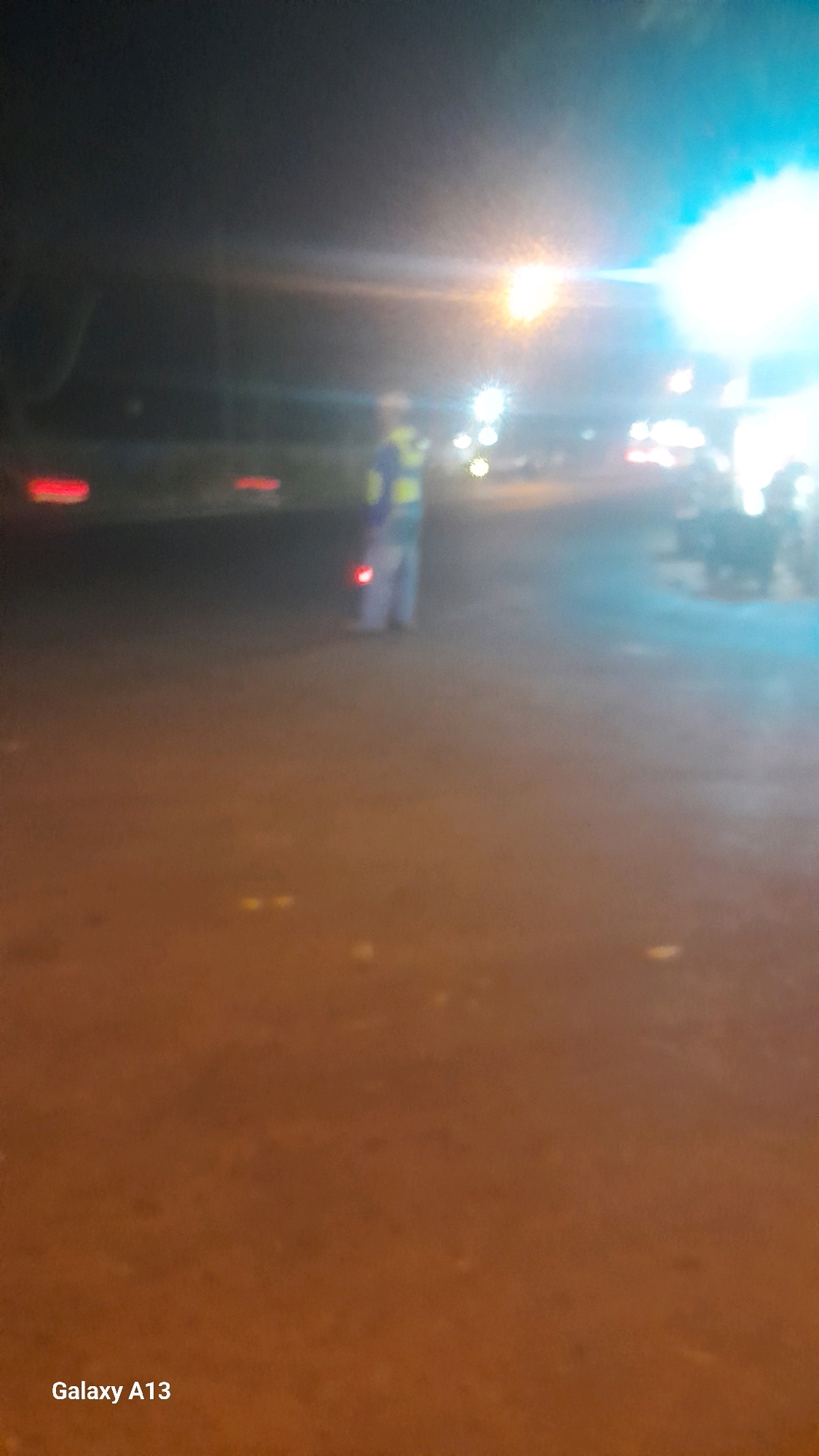 The image captures a nighttime street scene in Ngembalrejo, Indonesia. In the mid-ground, a person wearing a high-visibility vest, likely a road worker or security personnel, is visible. The individual appears to be standing, and their attire is predominantly yellow and blue. Behind this person, the scene is illuminated by bright artificial lights, possibly from street lamps or vehicle headlights, creating a hazy and somewhat blurred effect. Several red taillights from what appear to be vehicles are also visible on the left side of the image. The ground surface is dark and appears to be asphalt or a similar road material. The overall impression is of a dimly lit urban environment at night, possibly with traffic or ongoing work. The bottom left corner of the image displays the text "Galaxy A13", indicating the device used to capture the photo.