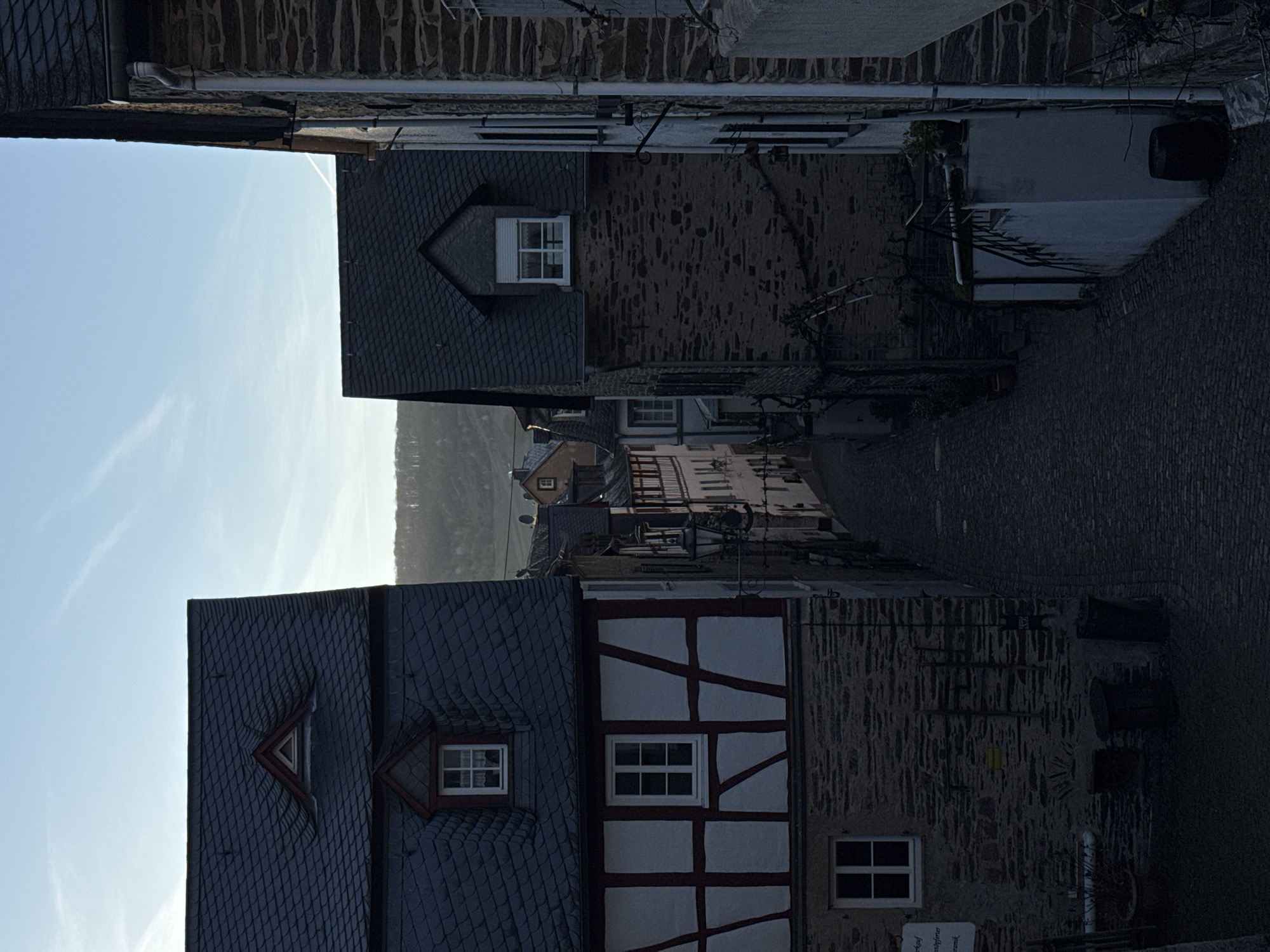 The image shows a view looking down a narrow street in Beilstein, Germany. The street is lined with old buildings, some with half-timbered construction and slate roofs. The sky is visible at the top of the frame. The street is paved with dark stone. No people are visible.