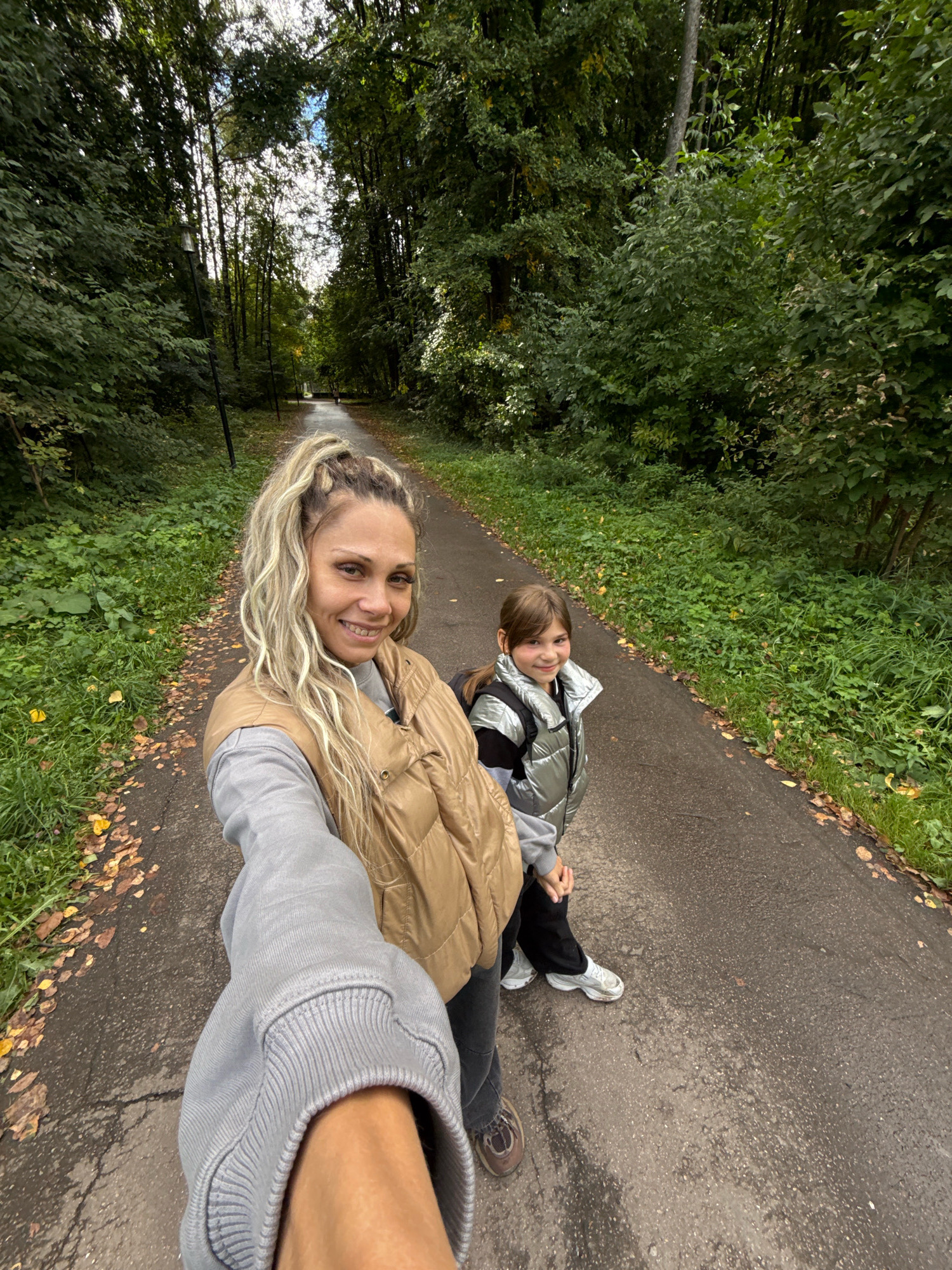 In Rusanovo, Russia, a woman with blonde hair in a ponytail and a young girl are walking on a paved path through a tree-lined park. The woman, in a tan puffer vest and grey sweatshirt, is holding the camera and smiling. The girl, wearing a silver puffer vest and black pants, is smiling and looking at the camera. Fallen leaves are scattered on the path and the surrounding grass. The trees are dense, with some sunlight filtering through.