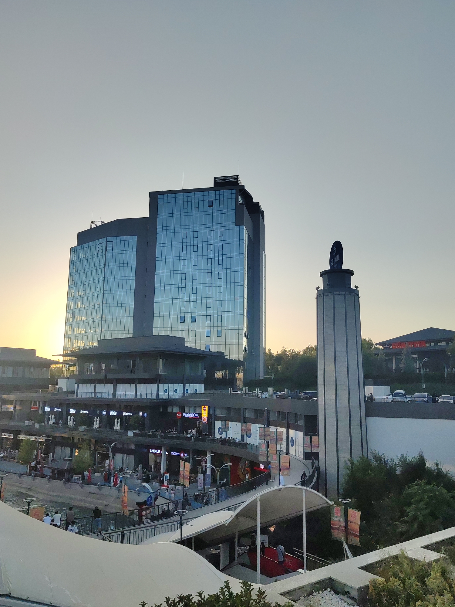 The image depicts a modern architectural complex in Tashkent, Uzbekistan, likely a commercial and entertainment district. In the background, a tall, glass-fronted building dominates the scene, reflecting the sky and the setting sun on its facade. To its right stands a distinctive, cylindrical tower topped with a circular sign that reads "SEOUL".
In the mid-ground, a multi-level structure with numerous shops and cafes is visible. Signs for businesses like "Gülfer Gelateria," "Mixue," and "VaponaM" are present. People are seen walking, sitting at tables, and interacting within this area, suggesting a lively atmosphere. Cars are parked in the background on an elevated roadway.
The foreground features an elevated walkway with a white, curved canopy, leading down towards a red escalator. More people are visible on the walkway and near the escalator. Lush green trees and foliage are scattered throughout the scene, adding a touch of nature to the urban environment. The sky is a soft, clear hue, indicating either early morning or late evening. The overall impression is one of a contemporary urban space designed for leisure and commerce.