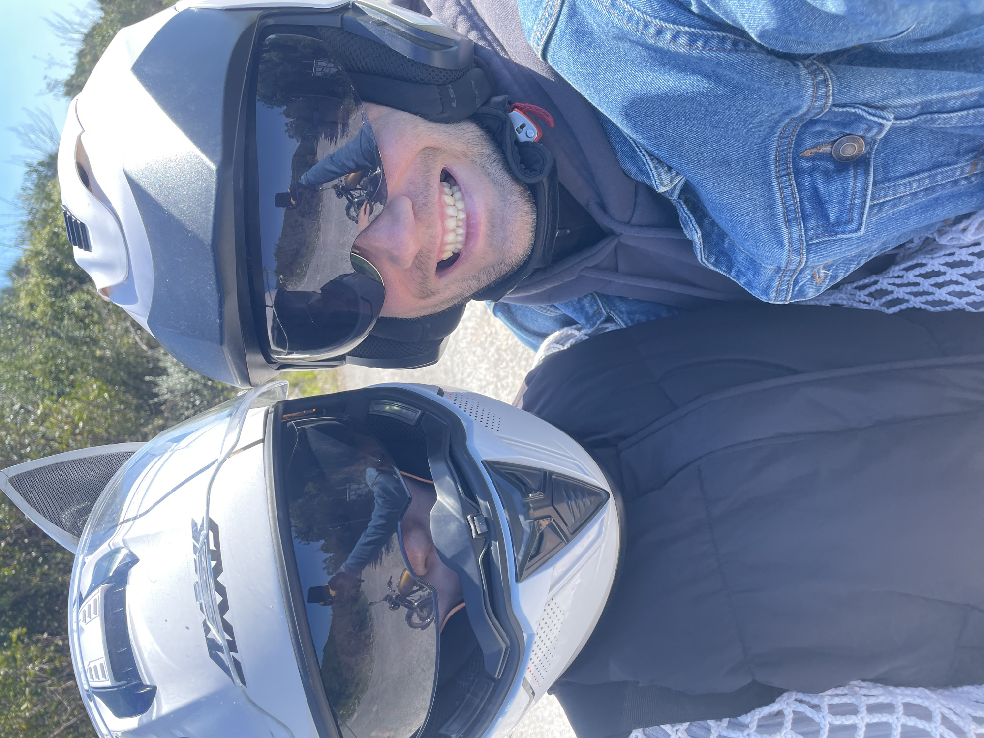 Two individuals are wearing helmets and possibly on a motorcycle, as suggested by the reflection in the visors showing a motorcycle and road. One person has a denim jacket and is smiling. The location appears to be outdoors on a sunny day, with greenery visible in the background, in what seems to be a rural or suburban area, possibly near Kubasi, Montenegro. The overall impression is that they are stopped on a road, likely taking a photo or break during a motorcycle ride.