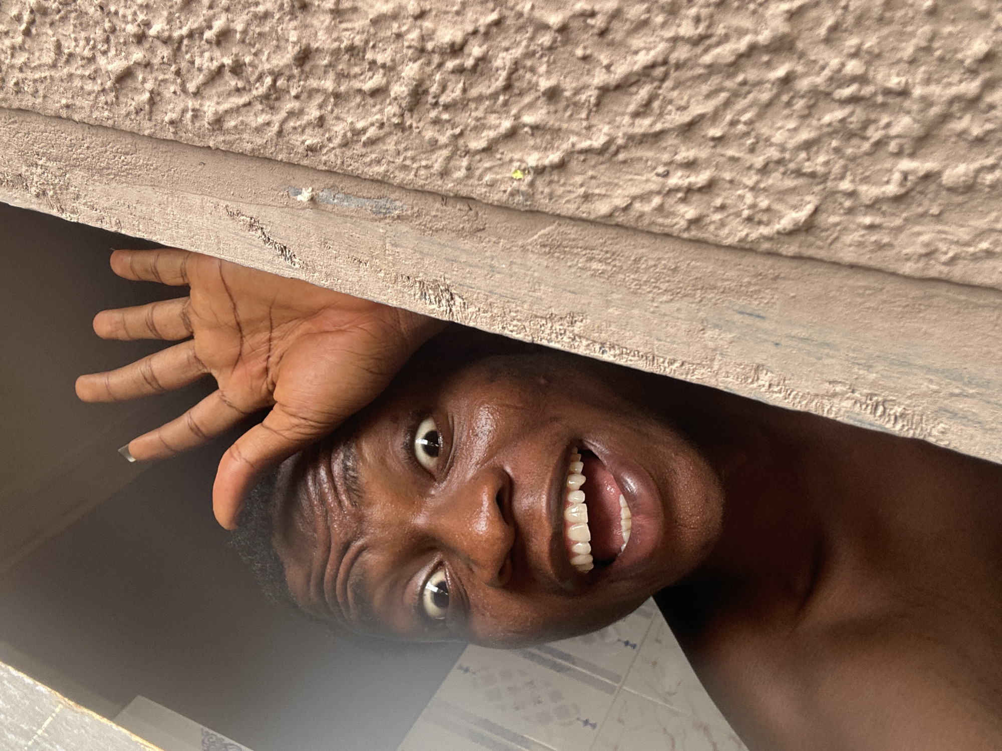 A close-up, slightly upward-angled shot shows a smiling dark-skinned person's face partially framed by a textured tan surface above and dark area on the left, suggesting the person is positioned looking down and inward from a space. The person's face shows visible sweat, wide eyes, and a wide smile revealing healthy teeth. A hand with fingers spread is positioned near the person's forehead, seemingly bracing against the surface above. The background features a glimpse of tile or marble-like material, suggesting a bathroom. The location is Unknown, Unknown.
