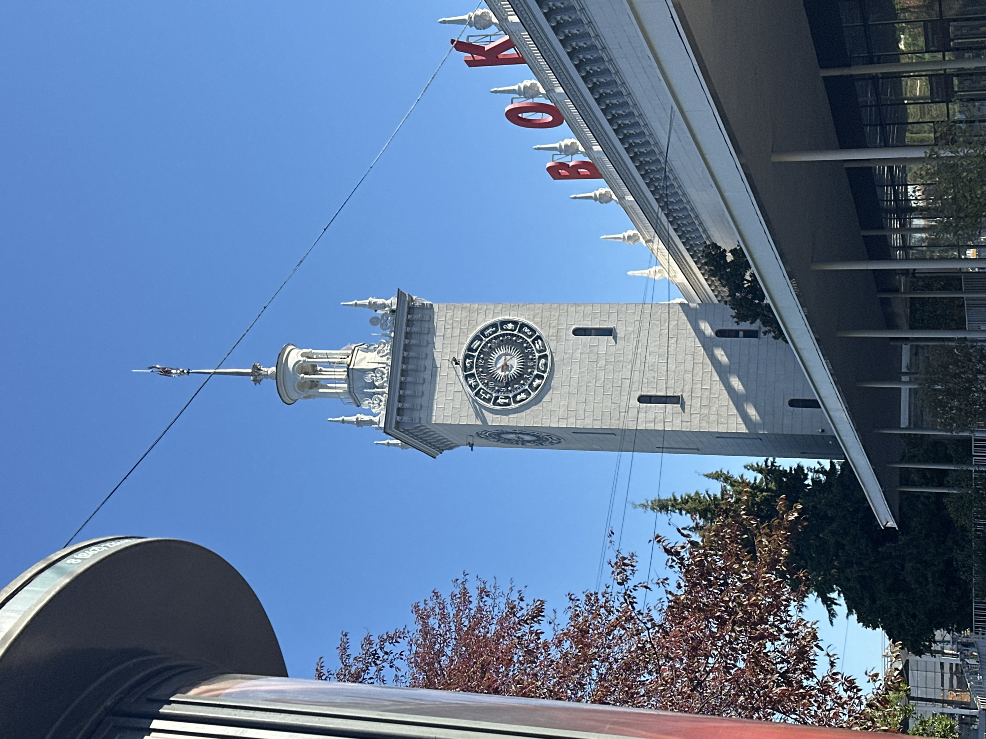 The image shows the Sochi railway station in Sochi, Russia, on a clear, sunny day. The main subject is the station's distinctive clock tower, a tall, grey, stone structure with architectural embellishments. The tower features a large, circular clock face with intricate markings and Roman numerals. Above the clock, the tower tapers to a spire topped with a weather vane.
To the right of the tower, the roofline of the station building is visible, adorned with decorative elements and topped with large, red, block letters spelling out "ВОКЗАЛ," which translates to "Station" in Russian. The building's facade is white and features a series of pointed spires along its crest.
In the foreground on the left, a portion of a cylindrical, metallic structure with vertical ribbed detailing is visible, partially obscuring a tree with red-tinged leaves. Behind the station building, evergreen trees are visible, suggesting a green, potentially park-like environment surrounding the station. The sky is a vibrant, cloudless blue. The scene depicts an architectural landmark with a surrounding urban or semi-urban environment.