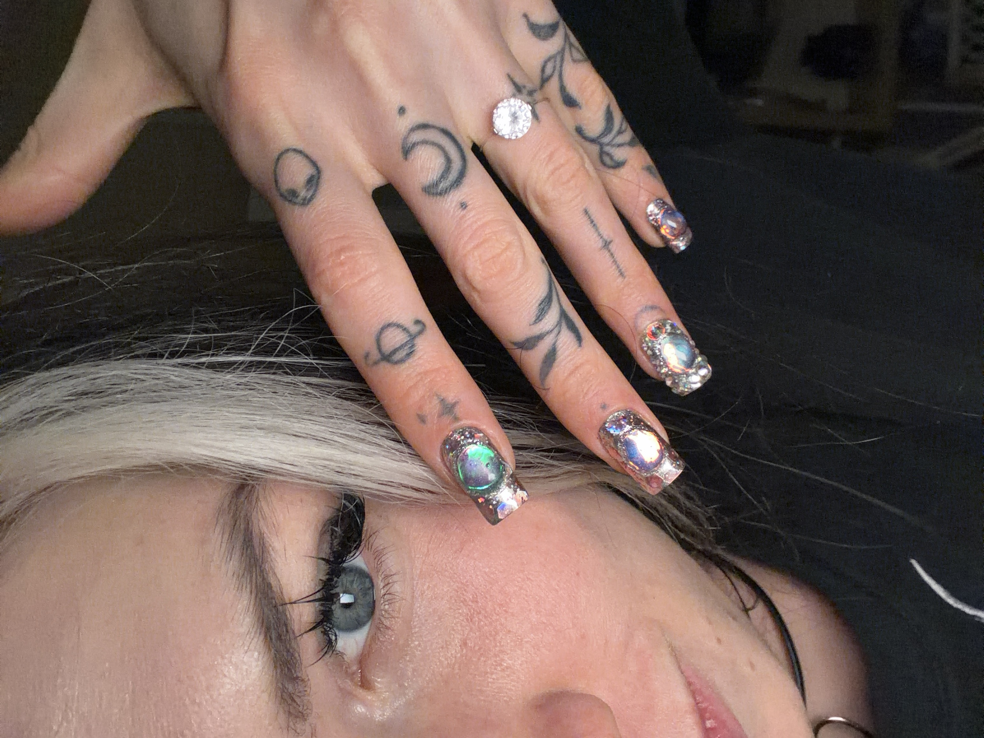A close-up, high-detail image captures a person's hand adorned with intricate tattoos and elaborate nail art, positioned above their face. The visible hand, likely the left, features several small, distinct tattoos on the fingers and knuckles, including an alien head, a crescent moon, a Saturn symbol, and various abstract or floral designs. A prominent ring with a large, round, faceted clear gemstone is worn on the ring finger. The fingernails are long and heavily embellished with numerous iridescent and clear gemstones, each featuring a large central, oval or round iridescent gem surrounded by smaller sparkling stones, against a metallic or glittery base.
Part of the person's face is visible in the lower half of the frame, showing a grey-blue eye with long, dark eyelashes, fair skin, and strands of light blonde or white hair blending into darker hair. The expression of the visible eye appears neutral. The background is dark and out of focus, suggesting either dark clothing or a dark surface. The image emphasizes personal style through detailed body art and accessories. There is no visible text.