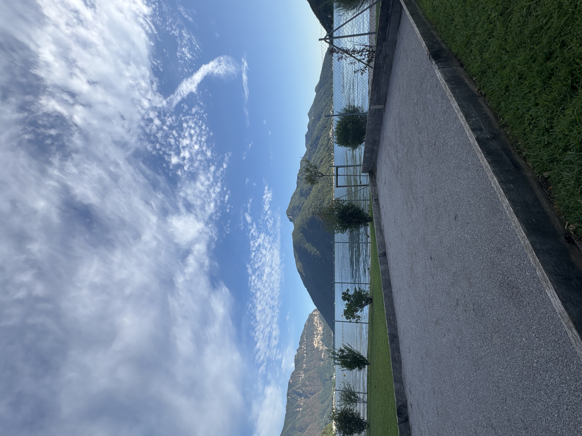 The image depicts a bocce ball court situated in Melide, Switzerland, with a serene lake and lush green mountains as its backdrop. The court, made of fine gravel, is enclosed by weathered wooden planks and bordered by a manicured lawn. A chain-link fence separates the court from the lake, beyond which several small, evenly spaced bushes line the shore. The lake itself is calm, reflecting the clear blue sky, which is adorned with wispy white clouds. In the distance, the majestic mountains, covered in dense green foliage, rise sharply from the lake's edge, with a hint of a small town visible nestled on their slopes. The lighting suggests it is either early morning or late afternoon, casting a soft glow over the entire scene. No people are present in the image.