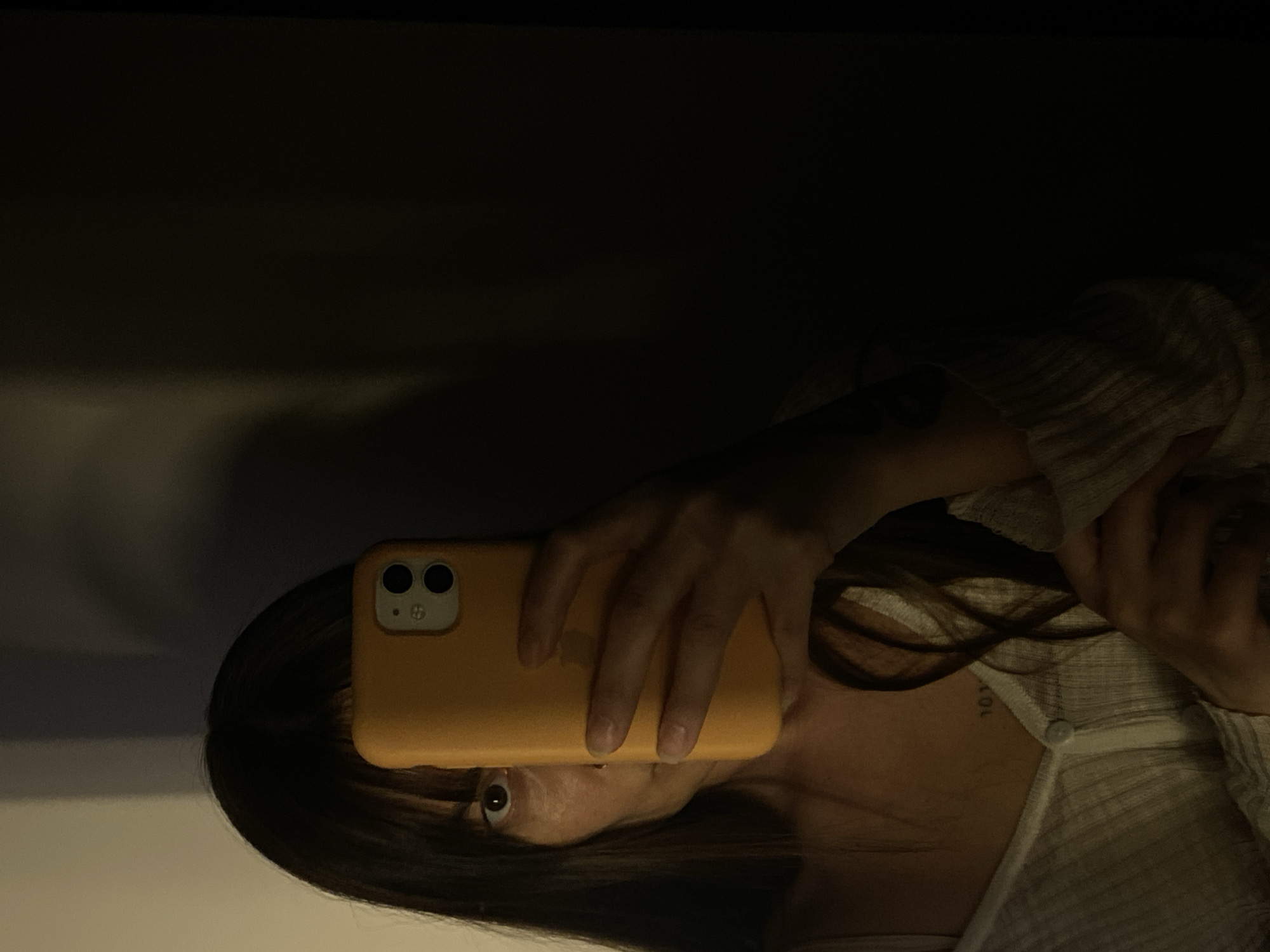 A person is holding an orange iPhone, likely taking a selfie. The person has dark hair with bangs, and is wearing a light-colored, buttoned shirt. There's a tattoo visible on their chest, above the buttoned shirt. The background is dark and blurred, potentially a wall or an undefined indoor space.
There is no explicit information about the location of this image being in Votkinsk, Russia. This cannot be inferred from the visual data.