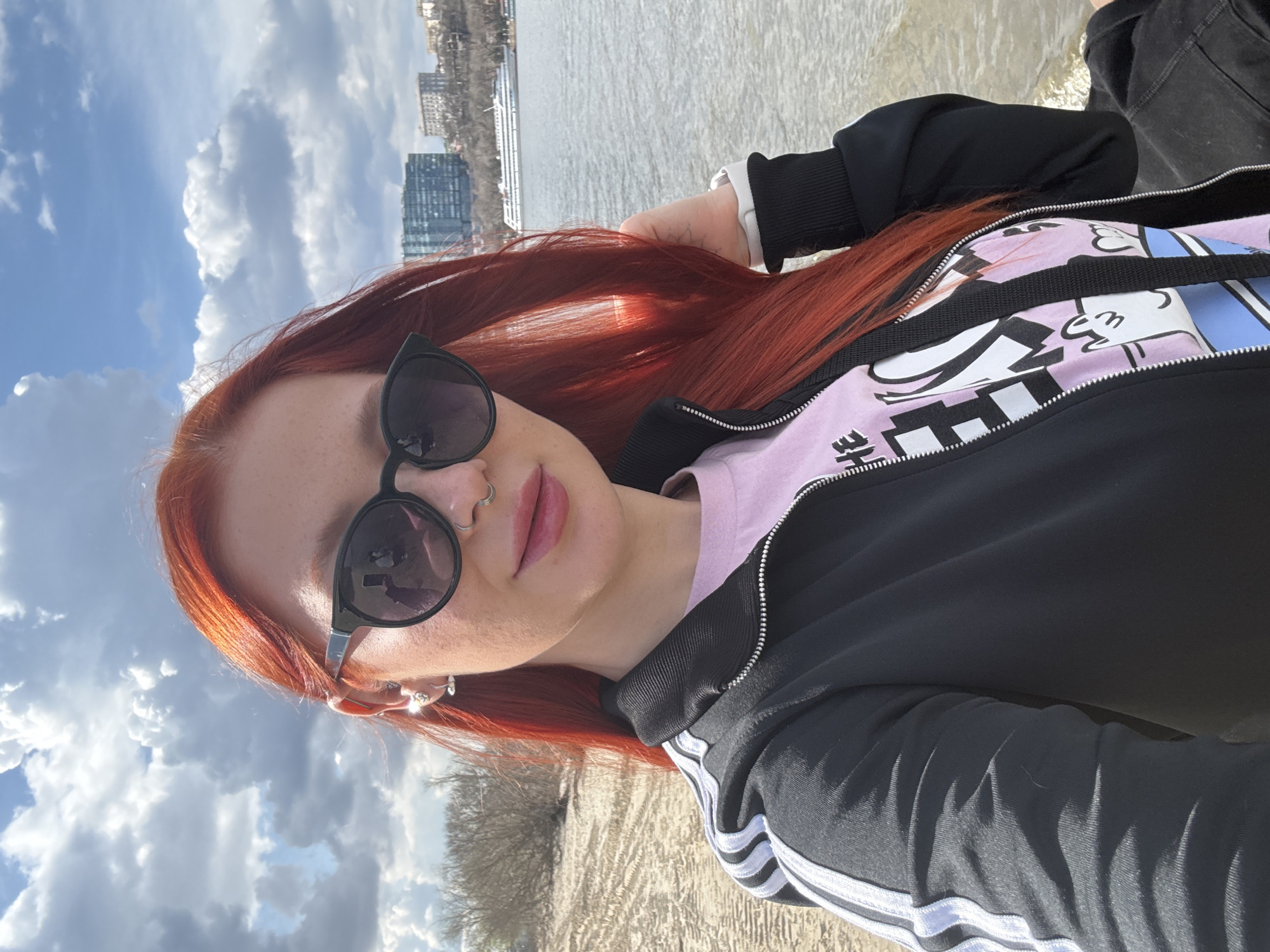 A selfie taken at a low angle depicts a woman with red hair wearing sunglasses and a black tracksuit jacket over a pink top. She has a nose ring and multiple earrings. The background reveals a city skyline across a body of water, likely the Don River, suggesting a location in Rostov-on-Don, Russia. The foreground includes sand or beach-like terrain, with a cloudy sky above. The image is rotated 90 degrees counterclockwise.