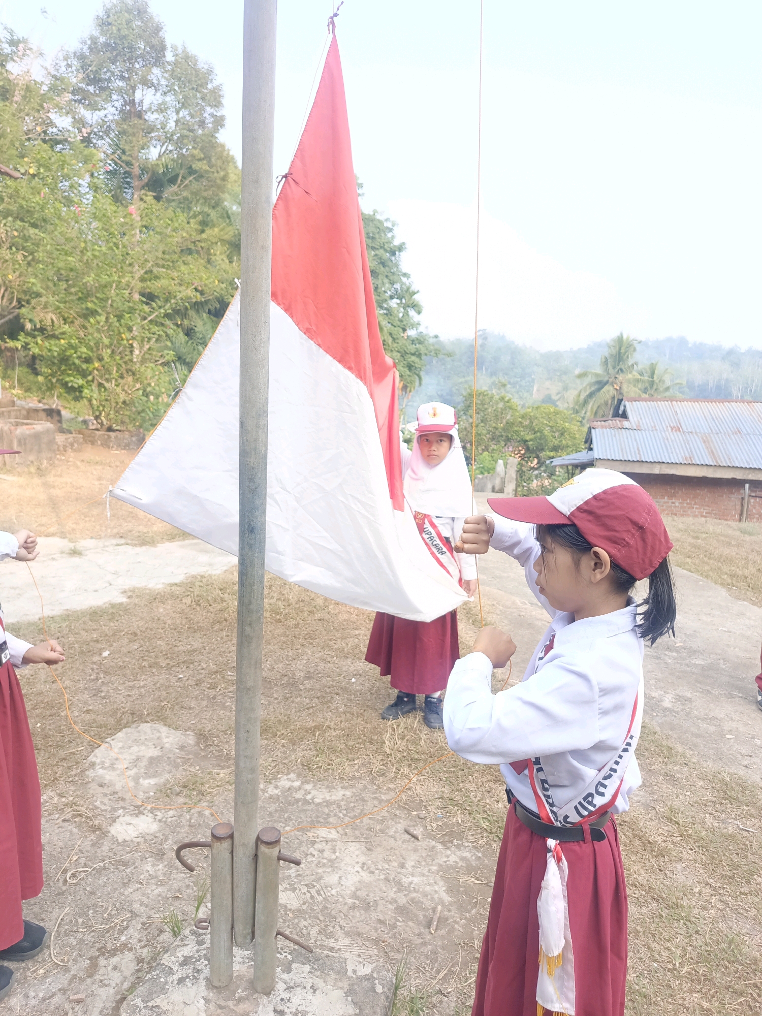 Two young girls in school uniforms are participating in a flag-raising ceremony. The girl in the foreground, wearing a red and white cap and a white shirt with a red sash, is pulling a rope to raise a red and white flag. The flag is partially unfurled and attached to a flagpole. Behind her, another girl, wearing a white hijab and a similar uniform, stands near the flagpole, holding the flag. The ceremony appears to be taking place outdoors in a rural setting, with trees, a building with a corrugated metal roof, and a hilly landscape visible in the background. The ground is dry and covered with grass and some scattered debris. The scene is set in Taba Tinggi, Indonesia.