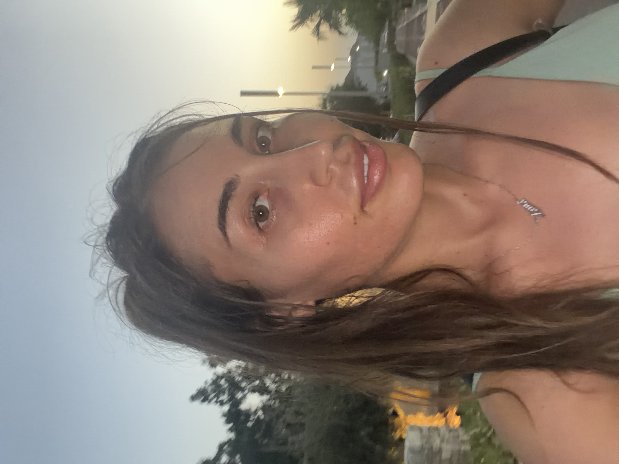 A medium-shot, selfie-style photograph features a young woman with brown hair pulled back, looking towards the camera. She has brown eyes, prominent cheekbones, and full lips. She is wearing a light green top and a silver necklace with the word "love" inscribed on it. Her hair is loose and falls around her shoulders. The background suggests an outdoor setting at dusk, with a soft, hazy sky. In the distance, trees, streetlights, and buildings are visible. The overall lighting is warm and atmospheric, indicative of a sunset or twilight hour. The location is Pervolia, Cyprus, a coastal area known for its beaches and relaxed atmosphere. The woman appears to be in good health and spirits.