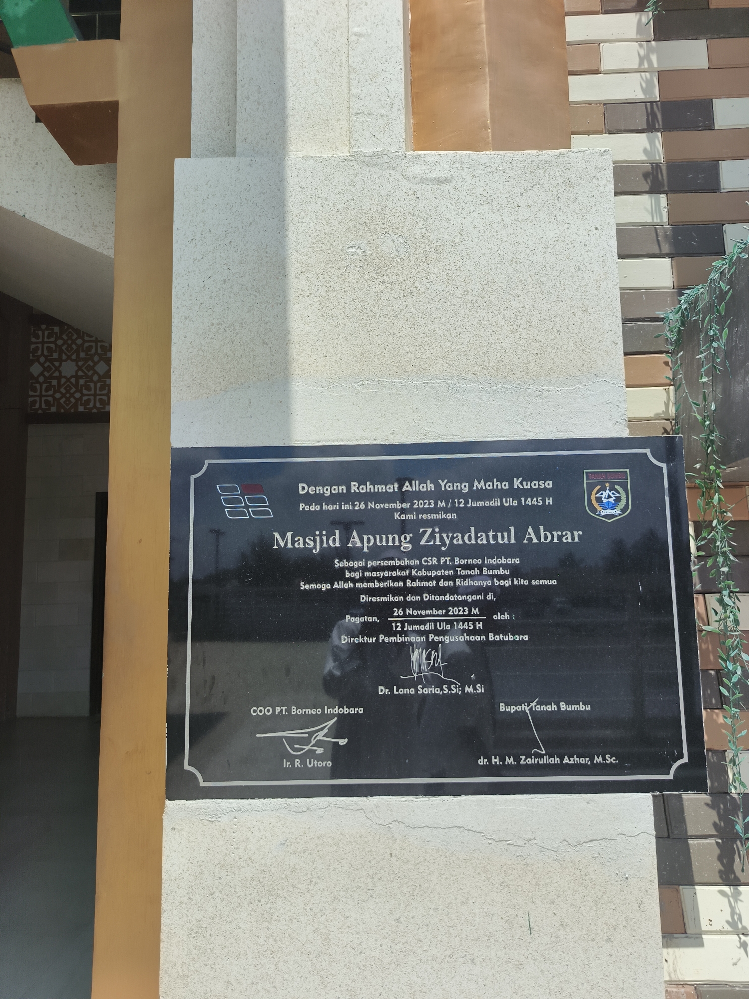 The image shows a plaque mounted on a textured concrete wall. The plaque is black with white lettering and contains text in Indonesian. At the top of the plaque, there is a logo with the text "BPNB" or a similar acronym and a stylized image. Below this, the text reads "Dengan Rahmat Allah Yang Maha Kuasa Pada hari ini 26 November 2023 M / 12 Jumadil Ula 1445 H Kami resmikan Masjid Apung Ziyadatul Abrar Sebagai persembahan CSR PT. Borneo Indobara bagi masyarakat Kabupaten Tanah Bumbu Semoga Allah memberikan Rahmat dan Ridhonya bagi kita semua Diresmikan dan Ditandatangani di, Pagatan, 26 November 2023 M oleh: 12 Jumadil Ula 1445 H Direktur Pembinaan Pengusahaan Batubara COO PT. Borneo Indobara Ir. R. Utoro Dr. Lana Saria,S.Si; M.Si Bupati Tanah Bumbu dr. H. M. Zairullah Azhar, M.Sc." The plaque is positioned in front of a building with a multi-colored brick facade and some green decorative foliage hanging from the right side. The location is identified as "Pagatan" in the text on the plaque, and a search indicates that Pagatan is a sub-district in the Tanah Bumbu Regency, South Kalimantan, Indonesia, and not in the city of Beringin.