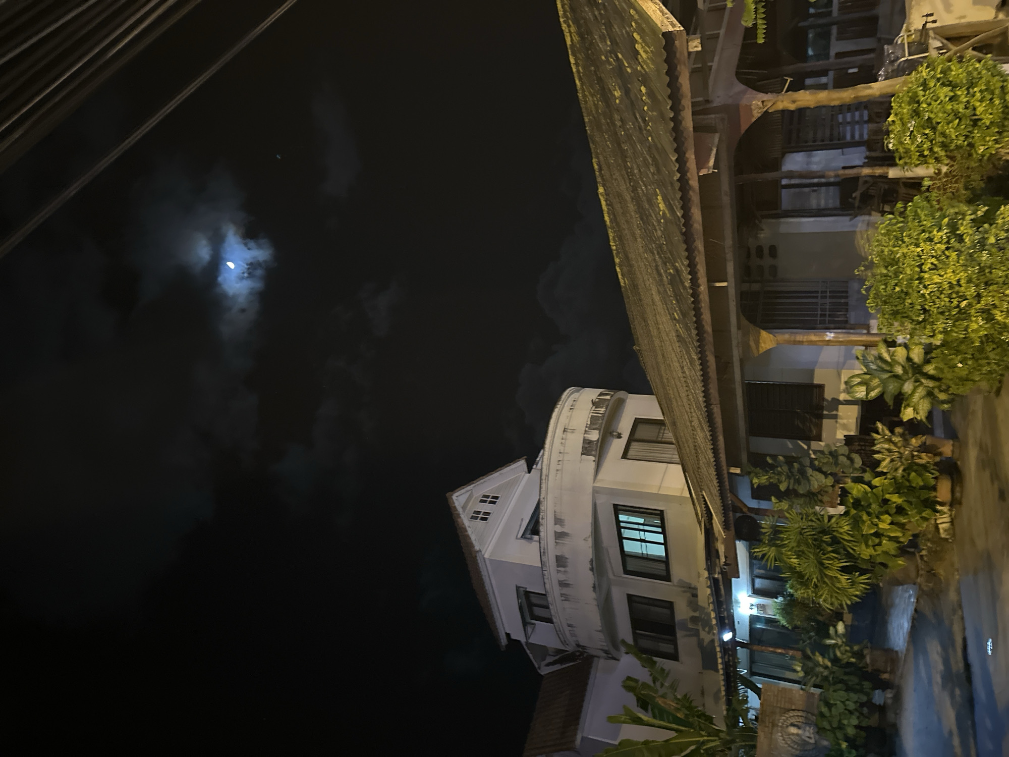 This media file depicts an outdoor scene at night, likely in Baan Mae Nam, Thailand. The image is oriented vertically, with the ground appearing at the top and the sky at the bottom.
The primary subjects are a building and the night sky. The building is a multi-story structure with white walls and dark-framed windows. It has a distinctive curved balcony or architectural feature. In front of the building, there is dense foliage, including trees and plants, illuminated by ambient light. A corrugated metal roof extends from a structure to the right, partially obscuring the view of the building and sky. Above the building, the night sky is dark, with clouds partially obscuring a bright, crescent moon. Electrical wires are visible in the upper left corner of the frame.
The scene captures a quiet, nighttime atmosphere. There are no people visible, and no specific activity or event is taking place. The lighting suggests it is nighttime, possibly late evening or early morning, and the presence of clouds indicates variable weather conditions, though no rain is apparent. The architectural style and vegetation provide some local context. The overall mood is serene and somewhat mysterious due to the low light conditions. There is no visible text in the image.