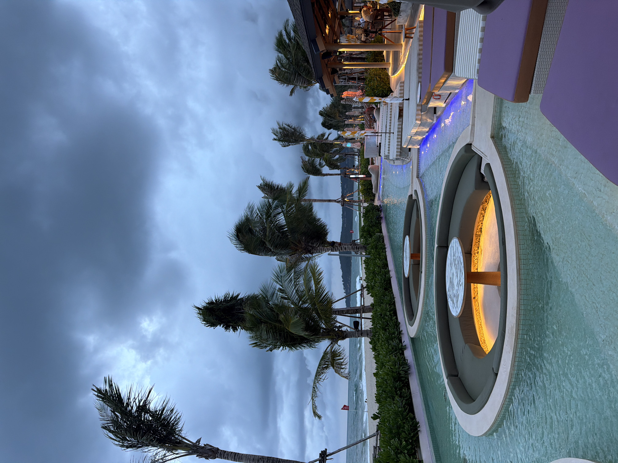 The image shows a scene at Choeng Thale, Thailand, at dusk or twilight. The sky is heavily overcast with dark, ominous clouds. Several palm trees line the beach, their fronds blowing in what appears to be a strong wind. The ocean is visible, with waves breaking on the sandy shore. A row of circular lounge seating areas is partially submerged in a shallow pool of water. These seating areas have tables with lit undersides. Further back, there is a building with an open-air dining area where people are seated. White lounge chairs are visible along the edge of the pool, and a small red flag is planted on the beach.