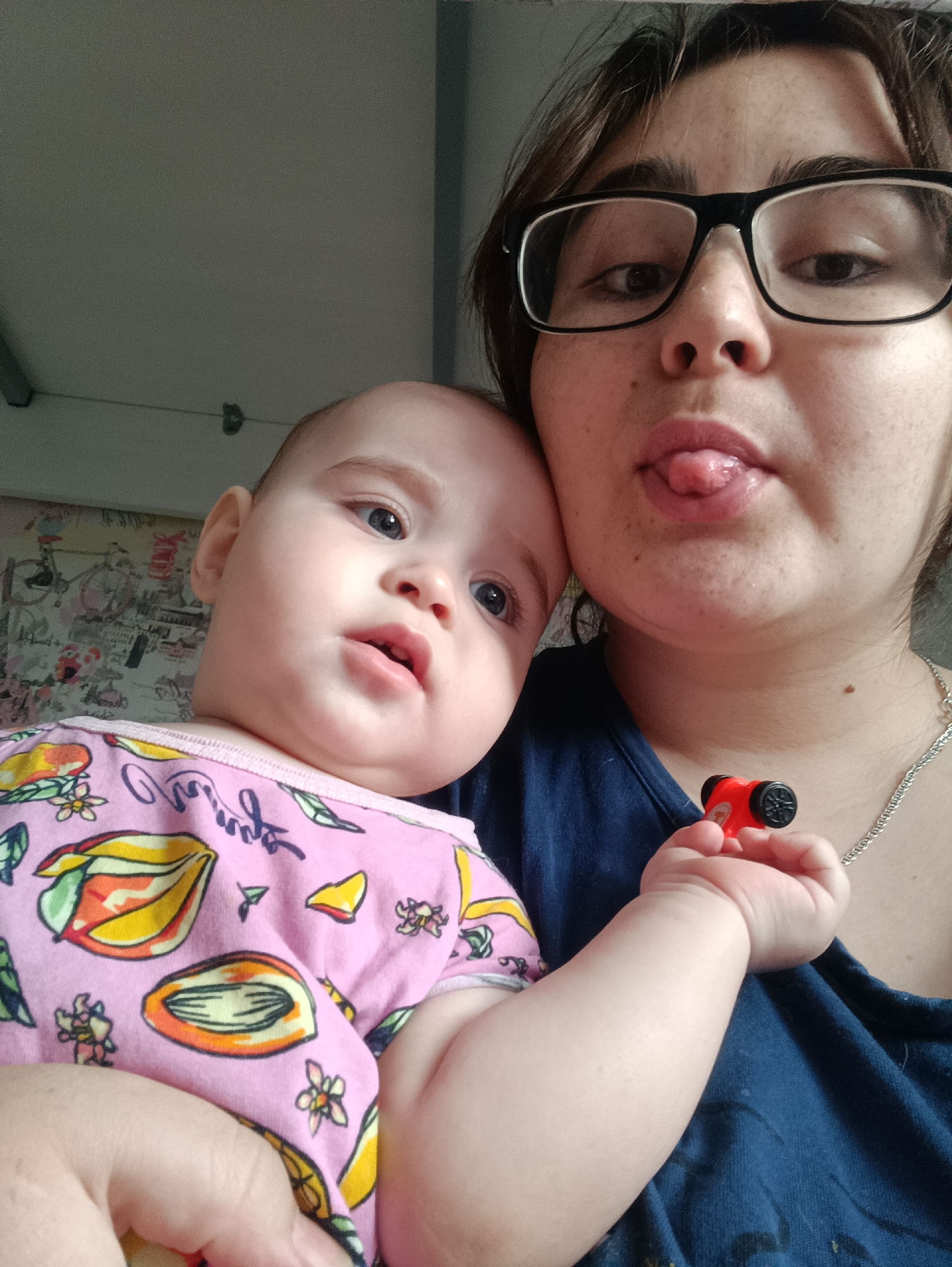 A close-up, high-angle selfie depicts a woman holding a baby. The woman, positioned on the right side of the frame, has brown hair, wears black-framed glasses, and a dark blue top. She is playfully sticking her tongue out towards the viewer. A silver chain necklace is visible around her neck, and she holds a small, red toy car with black wheels in her left hand, which is visible in the lower right corner.
The baby is situated on the left side of the image, featuring fair skin and light-colored eyes, gazing calmly towards the right. The baby is dressed in a pink long-sleeved outfit adorned with colorful illustrations of papayas, other fruits, flowers, and leaves. Some cursive text is also part of the pattern on the outfit, though it is not clearly legible. The baby's small, plump arm and hand are visible in the foreground.
The background behind the baby features a section of light-colored wallpaper with intricate black and white sketches of various elements, including cityscapes and bicycles. Above the baby and partially above the woman, a plain light-grey ceiling or wall is visible, along with some indistinct ceiling fixtures. The lighting appears natural and soft, suggesting an indoor setting during daylight hours. The image captures a tender and playful moment between the two individuals.