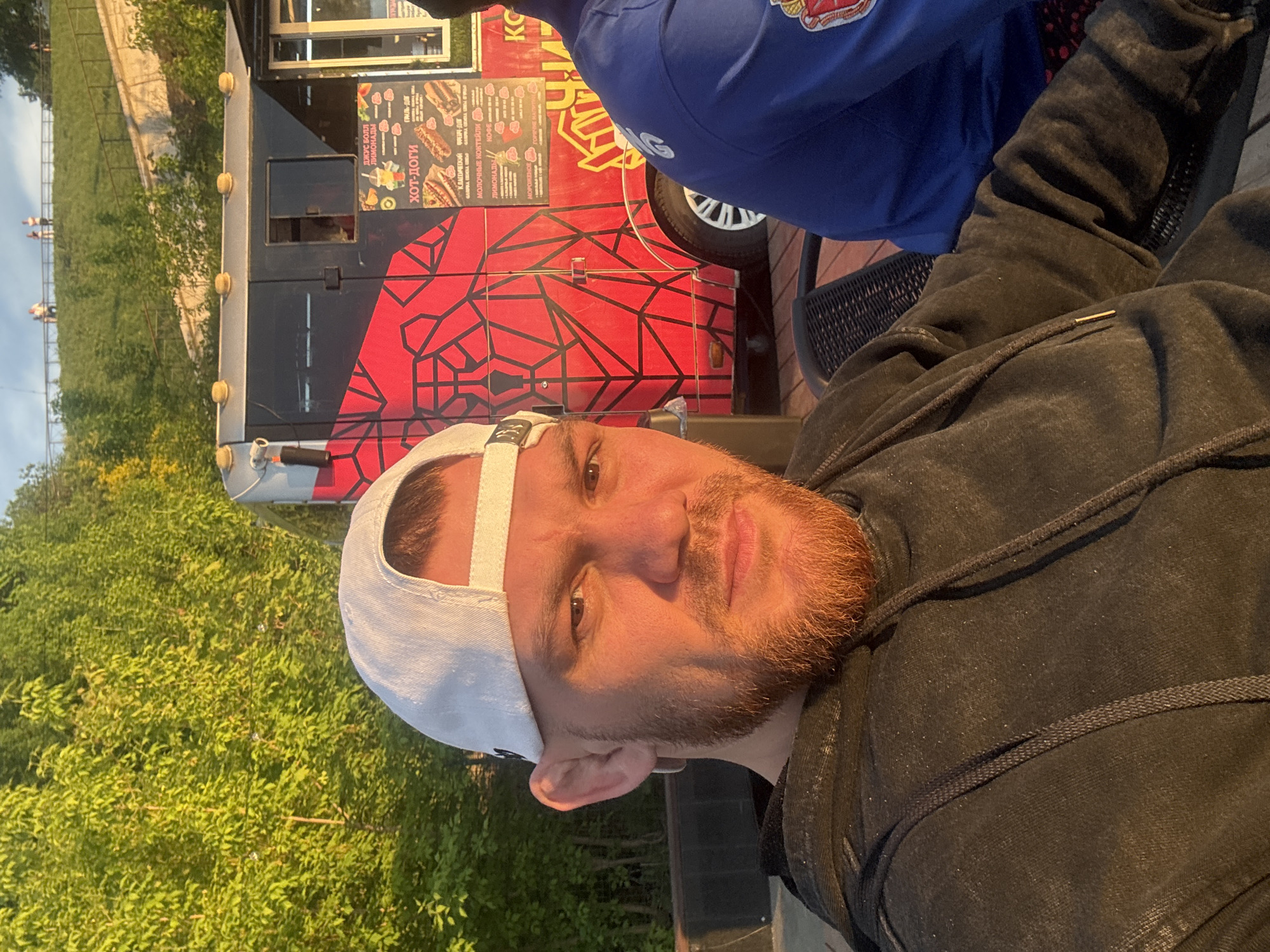 The image shows a selfie taken by a man with a fair complexion and a red beard. He is wearing a white baseball cap and a dark hoodie with a drawstring. Beside him is a food truck advertising "juice ball lemonades" and "hot dogs" in Russian. The food truck also lists "classic" and "brand" options and "milk shakes". The food truck is likely operating in Perm, Russia, given the Cyrillic lettering. Beyond the truck, there are trees and a distant embankment.