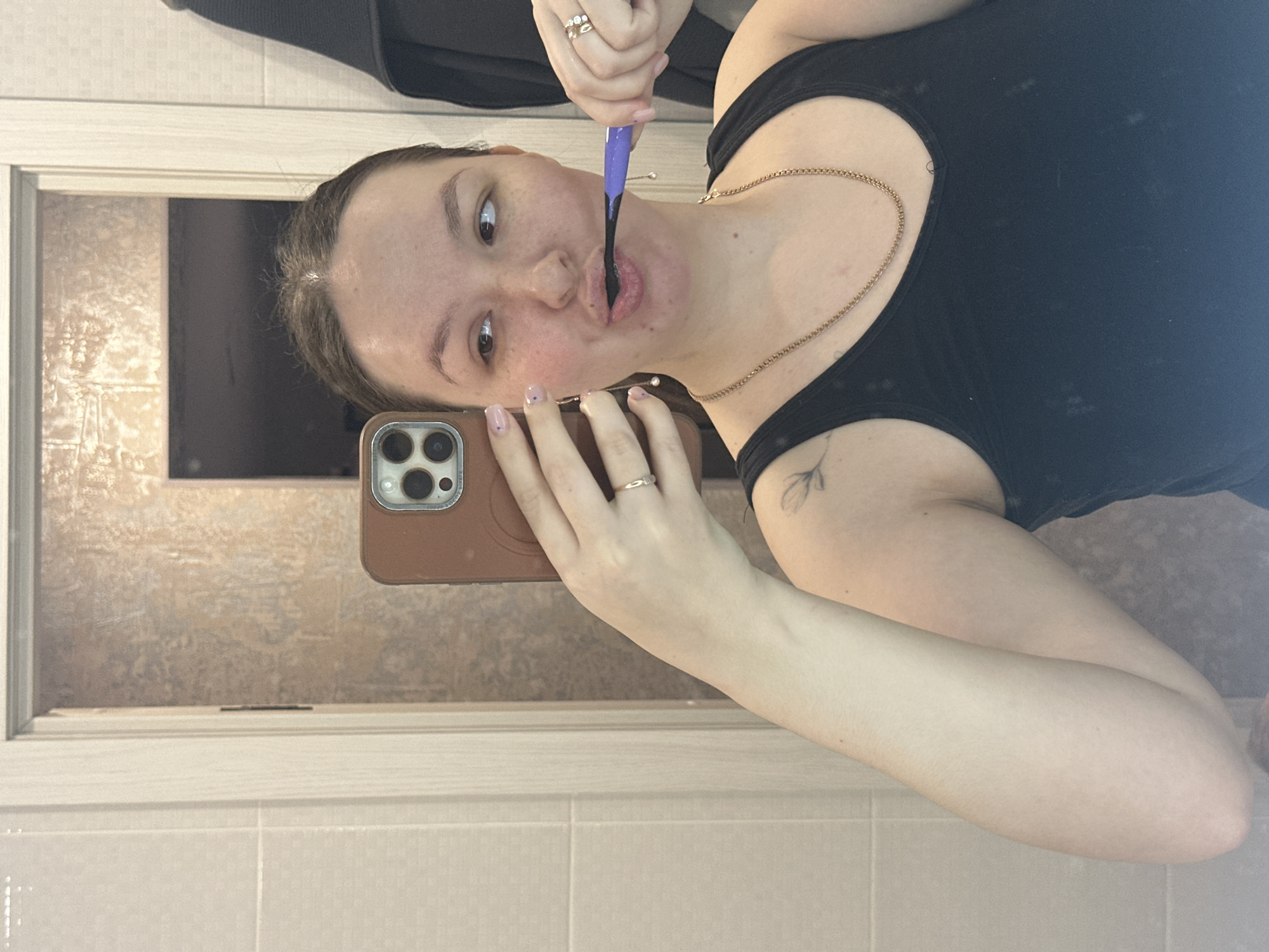 The image captures a young adult female in a bathroom setting within the city of Chaikovsky, Russia, engaged in a mirror selfie while brushing her teeth. The subject has pulled-back brown hair and a natural complexion. She is wearing a black tank top and a gold-colored chain necklace. On her left hand, she displays a gold-colored ring on her ring finger, and her fingernails are manicured with light pink polish featuring small blue dots. A stylized floral tattoo is visible on her left shoulder. She holds a purple-handled toothbrush with black bristles in her right hand, actively brushing her teeth with the brush inside her mouth, and her gaze is directed to her right. In her left hand, she holds a smartphone with a brown case, positioned to capture the reflection. The background, visible in the mirror, consists of light-colored, tiled walls on the left and a section of textured brown wall on the right. A white door frame is partially visible on the left, and a dark, possibly black, towel or garment hangs on a hook to the far right.