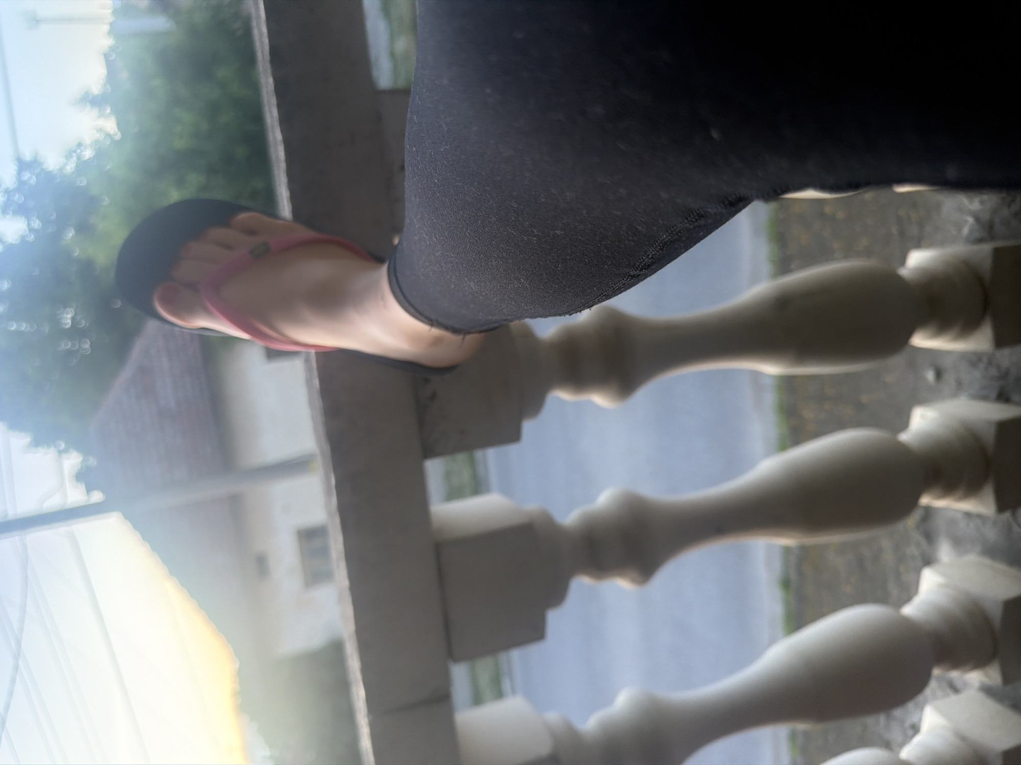 The image is rotated 90 degrees clockwise, depicting a close-up view of a person's lower leg and foot resting on a white balustrade. The person is wearing black leggings and a pink and black flip-flop with a small, indistinct green emblem on the strap.
The setting appears to be outdoors, likely on a balcony or porch overlooking a residential area. In the blurred background, there are green trees, a building with a light-colored wall and a reddish roof, and an expansive bright sky, suggesting clear, sunny weather. A soft, bright glow is visible in the lower left portion of the image, potentially indicating sunlight or a reflection.
The activity taking place is a person casually resting their foot on the railing. The image captures a moment of relaxation in an outdoor environment. There is no visible text in the image.