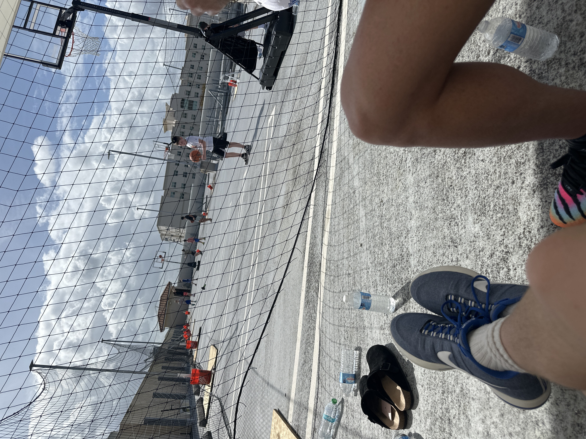The image presents a view of a basketball court in Aventura, Florida, United States. The foreground shows a person wearing blue sneakers and white socks. Near this person are a water bottle and a pair of black shoes.
In the background, a basketball game is in progress. The court is surrounded by a high, black net and orange cones are visible near the court boundaries. The sky above the court is partly cloudy. A building is visible near the edge of the court, partially obscured by the net.