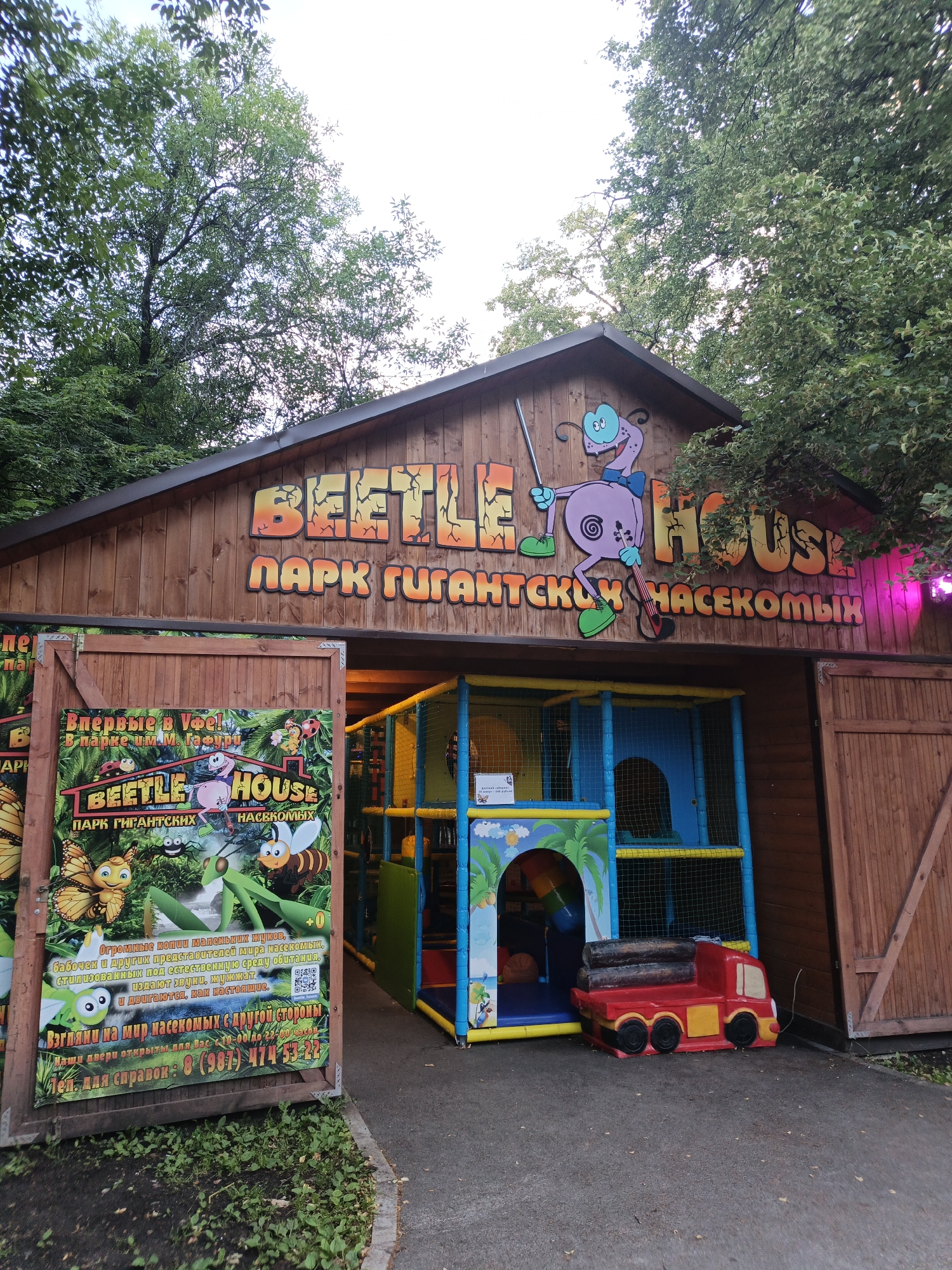 The image shows the entrance to "Beetle House," a park in Ufa, Russia, featuring giant insect exhibits and a children's play area. The building is made of wood and has a large sign with the park's name and a cartoon character. To the left of the entrance is a large promotional poster with images of various insects and information about the park. The play area is visible inside the building, with colorful slides and climbing structures. A red toy truck is placed on the ground in front of the play area. The scene is set outdoors, surrounded by trees, indicating it is likely a warm day.