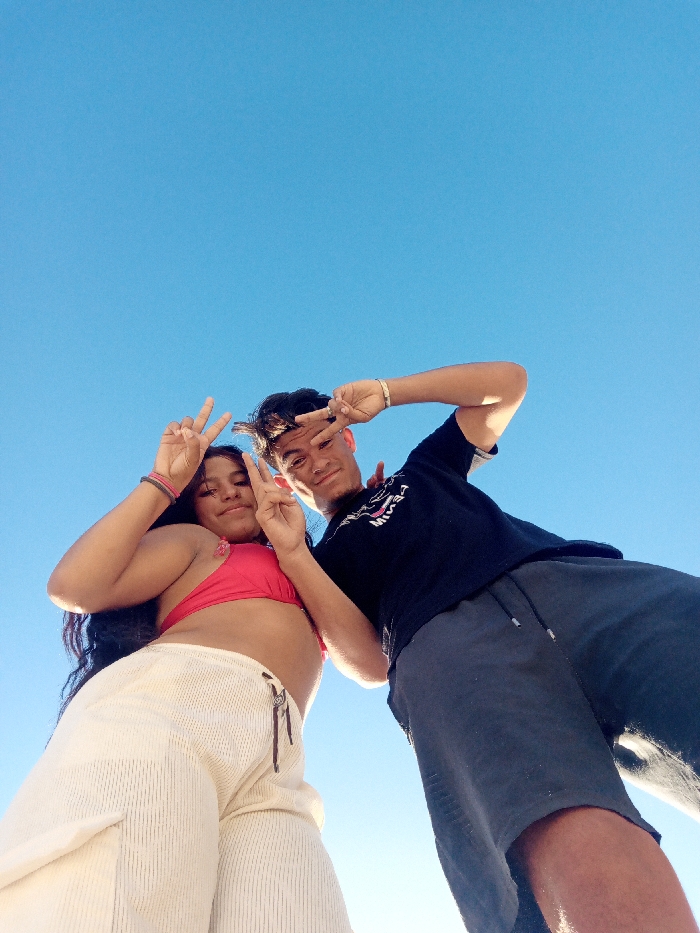 A couple poses against a bright blue sky. The woman, wearing a red bikini top and white pants, holds up a peace sign. Beside her, a man in a black t-shirt and gray shorts mirrors the gesture. The low-angle shot emphasizes the sky and their casual attire suggests a warm, outdoor setting.