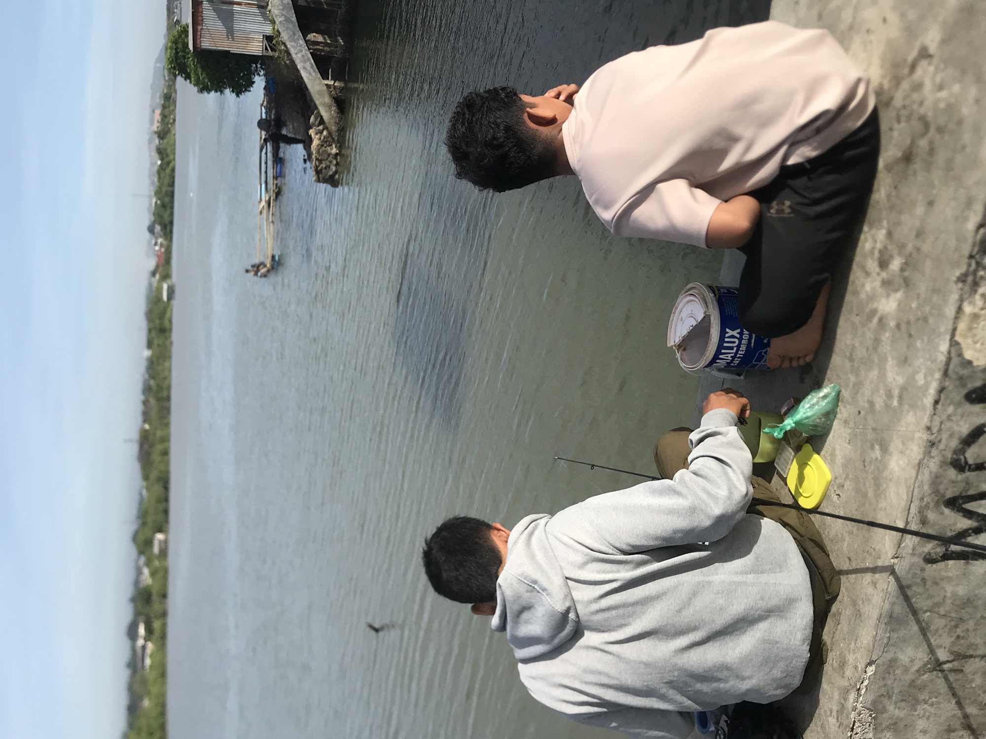 Two people are fishing from a concrete bank along a river in Lhokseumawe, Indonesia. One individual, wearing a light pink shirt and dark pants, kneels barefoot next to a blue paint bucket. The second individual, wearing a gray hooded sweatshirt and pants, holds a fishing rod. Fishing gear, including a green plastic bag and a yellow lid, lie on the concrete near the second person. In the background, a building on stilts stands over the water, with the opposite shore lined with trees. The sky is clear and light blue.