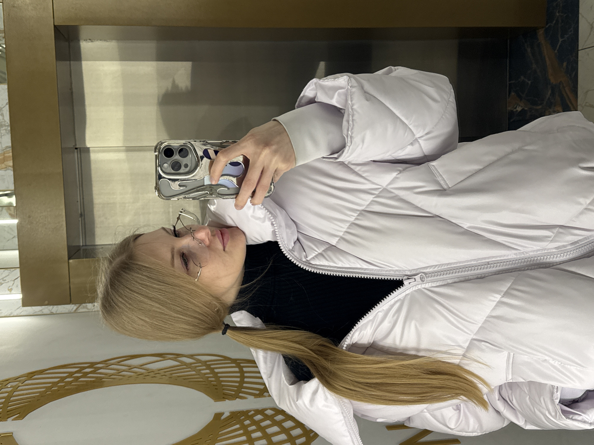 A blonde-haired woman with eyeglasses takes a selfie in an elevator with a marble and gold accent. She wears a black turtleneck under a white quilted jacket. The elevator appears to be in Perm, Russia, based on architectural styles, patterns, and overall design.
