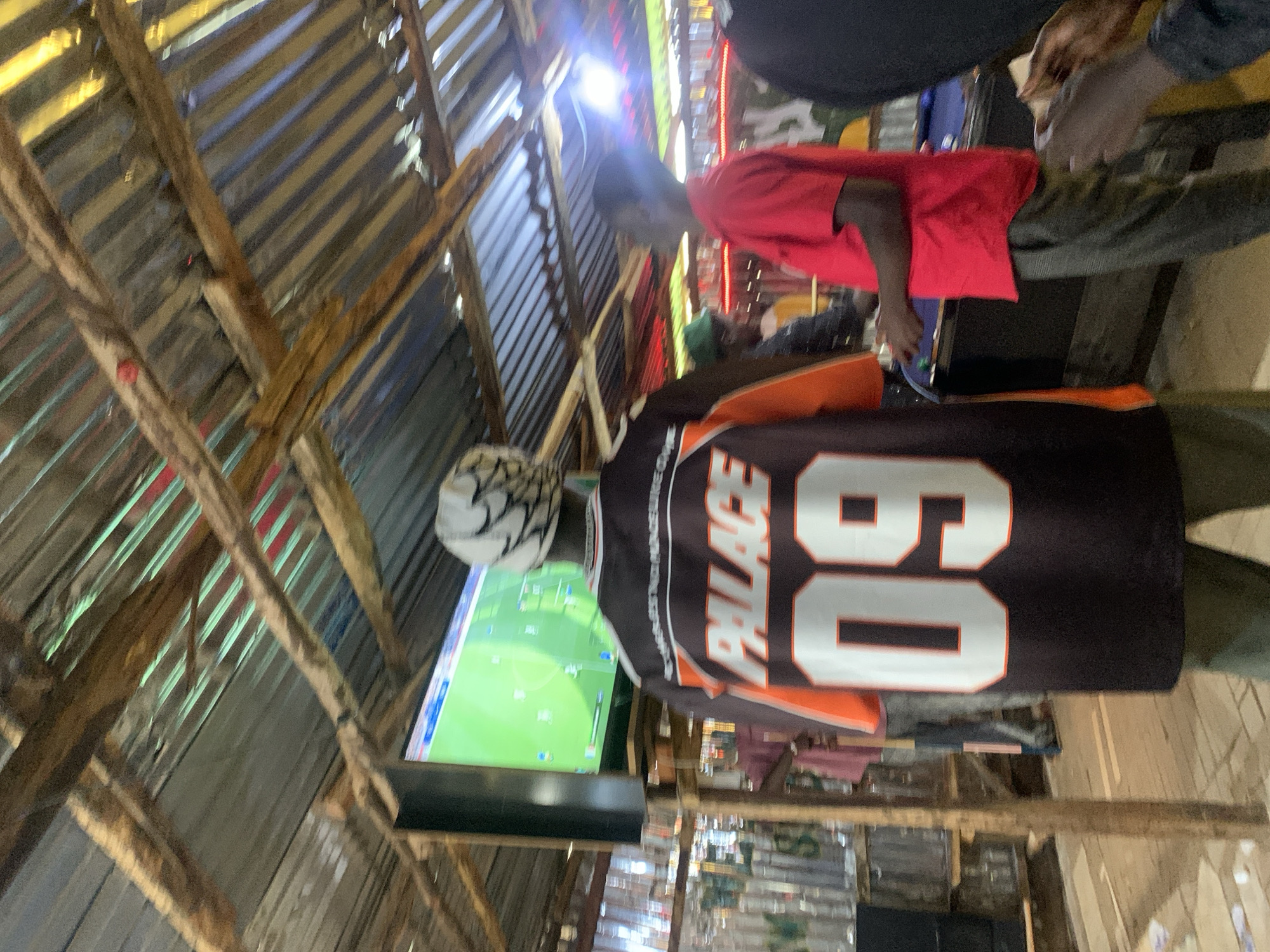 The image depicts an outdoor, open-air venue in Durumi, Nigeria, with a corrugated metal roof supported by rough-hewn wooden beams. In the foreground, a person wearing a black and orange jersey with "PALACE 09" prominently displayed is facing away from the camera, looking towards a large television screen. The screen displays a video game, likely a soccer simulation, with a green field and player icons. To the right, another individual in a red shirt and grey pants is visible, also appearing to be engaged with the activity, possibly playing a game on a pool table situated behind them. The environment suggests a casual gathering place, possibly a bar or entertainment spot, with shelves of glasses visible in the background and ambient lighting creating a lively atmosphere.
