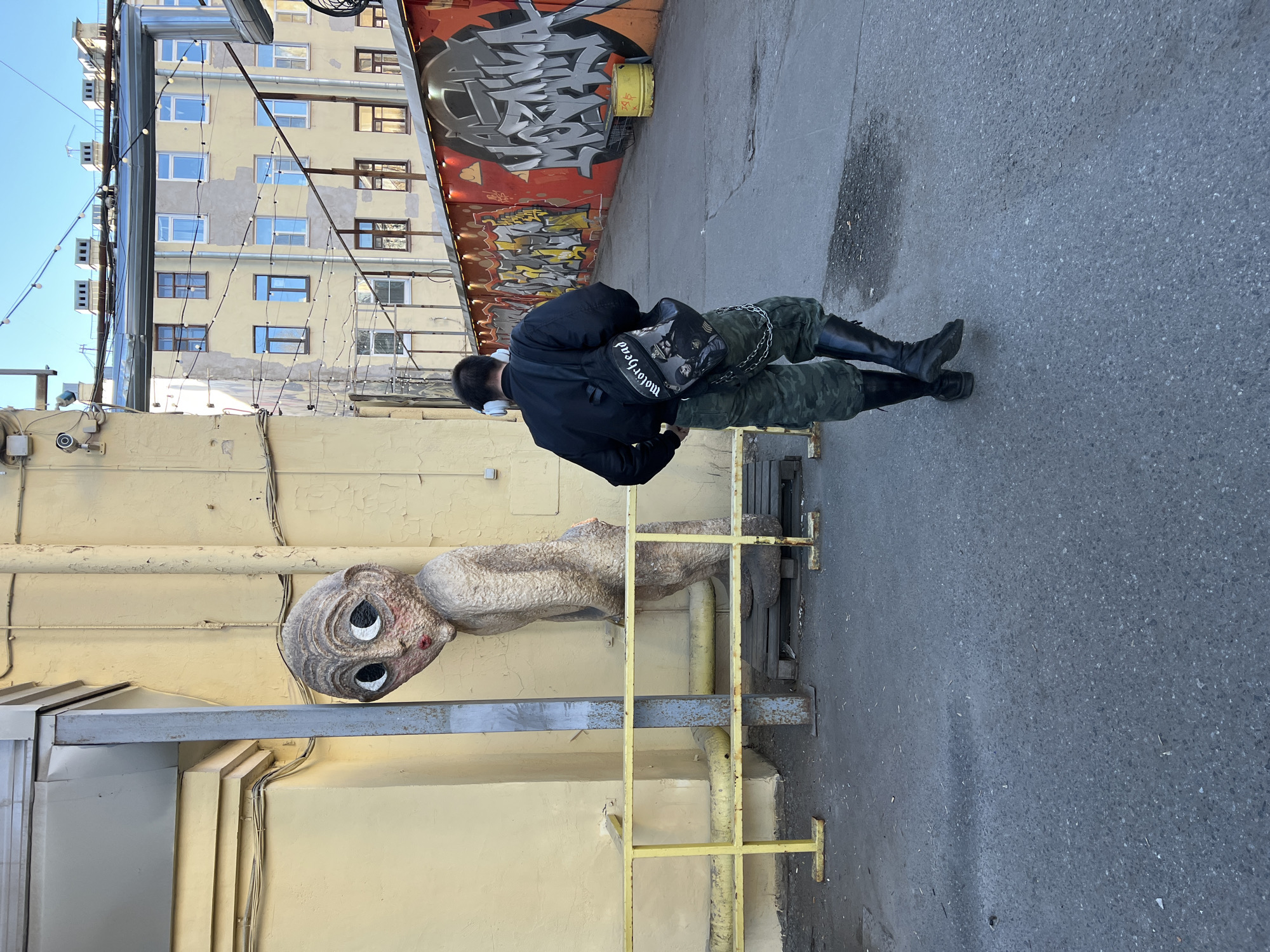 The image shows a person standing in an alley in Saint Petersburg, Russia. A sculpture of an alien head is attached to the wall on the left. The person is wearing a black jacket, camouflage pants, and black boots. They have a backpack on and are wearing headphones. The background includes a building with multiple windows and graffiti on a wall. A yellow ladder is positioned against the wall near the sculpture.