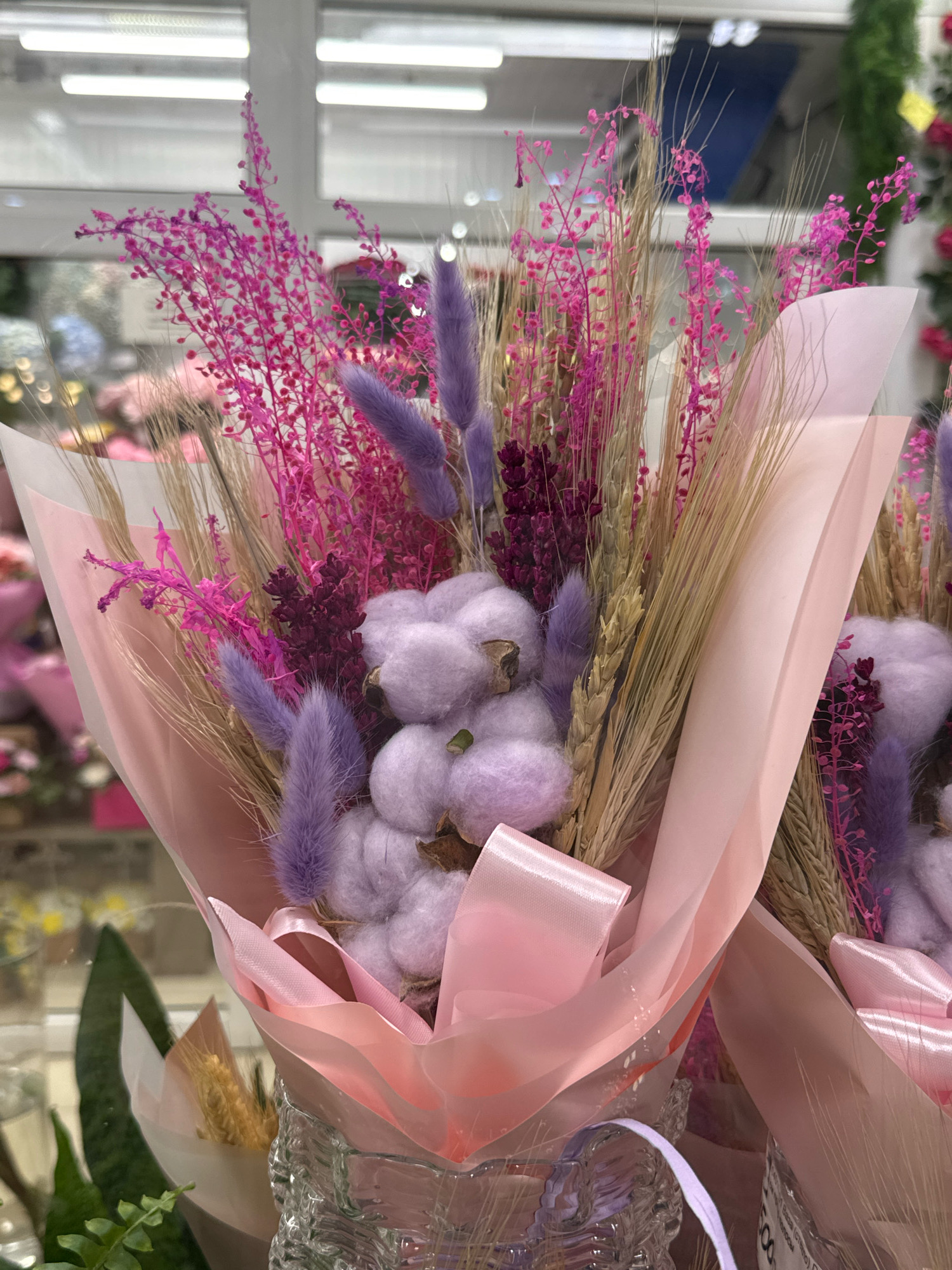 The image shows a close-up of two bouquets of dried flowers, likely displayed in a flower shop. The bouquets are predominantly pink and purple, with accents of wheat stalks and fluffy white cotton balls. They are wrapped in pale pink paper and tied with pink ribbons. The background is blurred but suggests a retail environment with bright fluorescent lighting and displays of other floral arrangements. One bouquet is more in focus than the other, showing intricate details of the dried flowers and their arrangement. The bouquet on the left is partially obscured by the bouquet on the right. A clear glass vase with a textured surface holds the bouquet on the left. The overall impression is one of delicate, dried floral artistry. The scene is set in an indoor commercial space.