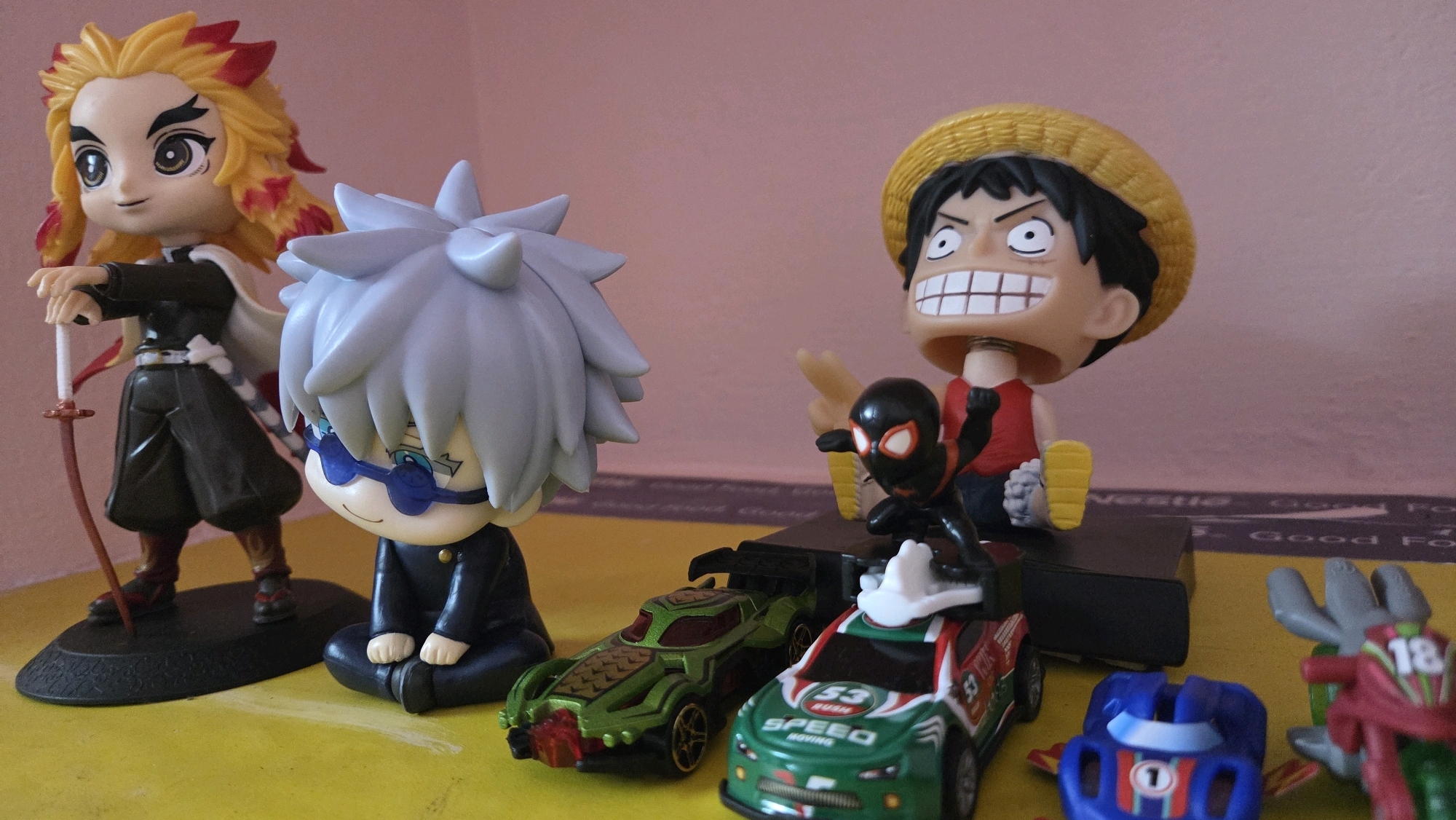 In a close-up, low-angle indoor shot, several anime-style figures and toy cars are arranged on a yellow surface against a soft pink background. On the left, a female anime figure with fiery red and yellow hair, wearing a dark uniform and a white cape, holds a sword. Next to her, a male anime figure with spiky light gray hair and blue glasses sits cross-legged in a dark uniform. To the right, a large anime figure with a yellow straw hat, red shirt, and wide grin is positioned behind a smaller figure dressed as Spider-Man in a black and red suit. In front of these figures are two toy race cars: a green one with gold accents and a red and green one with white and red lettering. A blue toy car with a "1" on it is visible in the foreground, along with a portion of a red and green toy motorcycle. The lighting is soft, creating a warm atmosphere. There is no indication of Chalakudy, India in the image.
