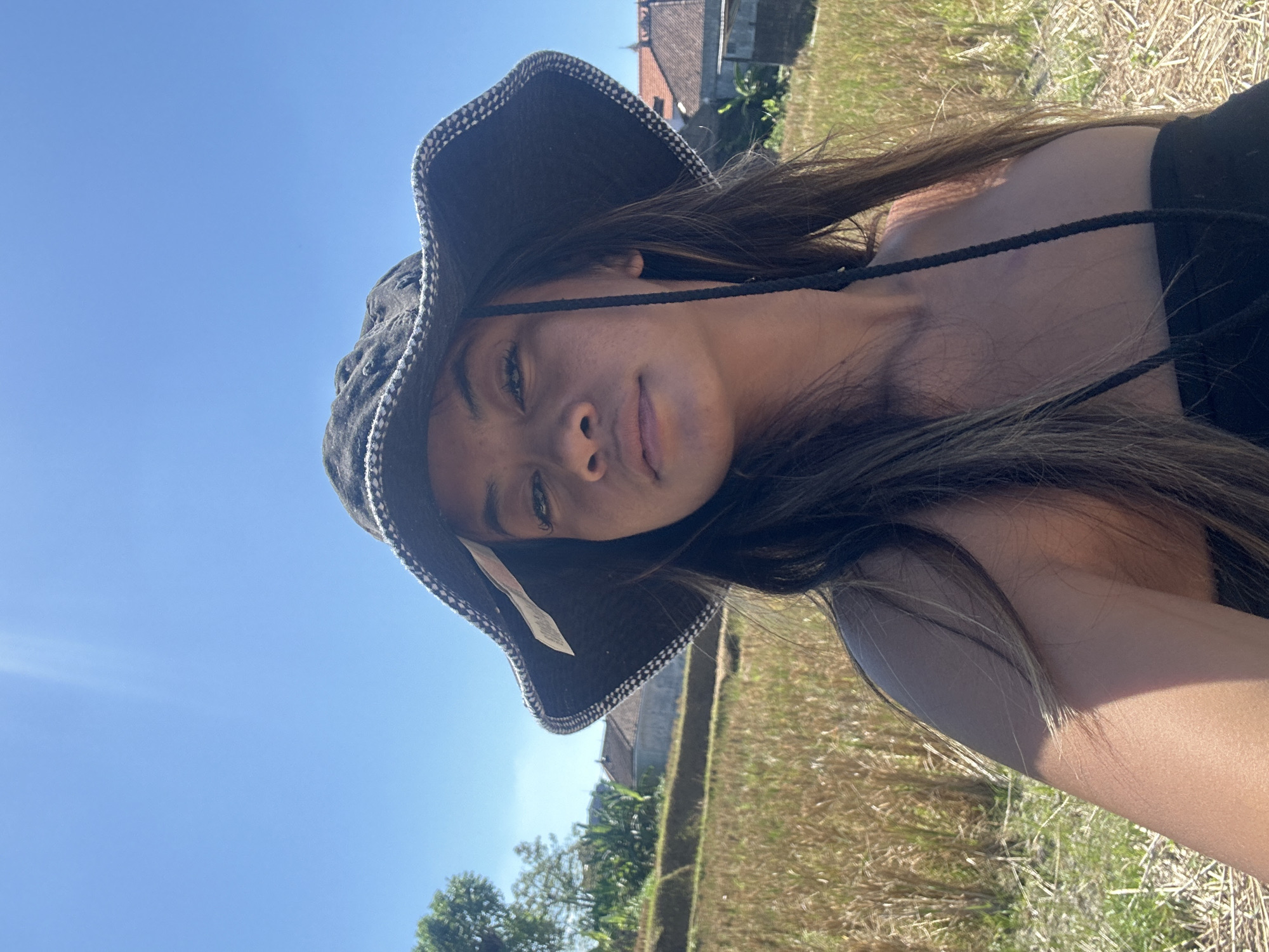 A young woman wearing a black wide-brimmed hat stands in a dry rice paddy field under a clear blue sky. The field is filled with golden-brown stubble, indicating a harvest has recently taken place. In the background, there are some traditional Balinese houses with terracotta roofs and lush green trees. The woman is facing the camera, looking directly at the lens with a neutral expression. Her long, dark hair cascades over her shoulders. The scene appears to be located in Sukawati, Indonesia, a region known for its rice terraces and traditional villages. The lighting suggests it is a bright, sunny day.