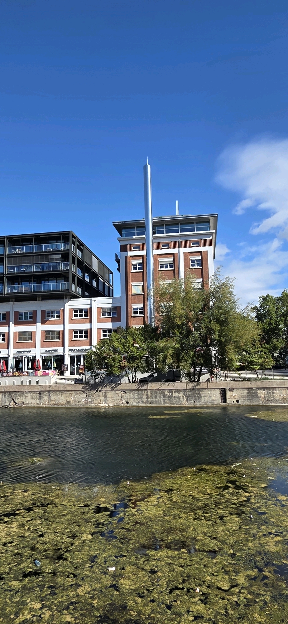 This image captures a canal in Strasbourg, France, with a modern building complex situated along its bank. The water in the foreground is covered with a thick layer of green algae and some scattered debris. Beyond the canal, a concrete retaining wall lines the bank, supporting a paved walkway. Behind the wall, a multi-story building with a brick facade and white window frames is visible, featuring a tall, slender metal chimney extending upwards. To the left of this building is a darker, more contemporary structure with multiple balconies. Trees with green foliage are interspersed along the bank, and the sky above is a clear blue with a few wispy white clouds. No people are visible in the scene.