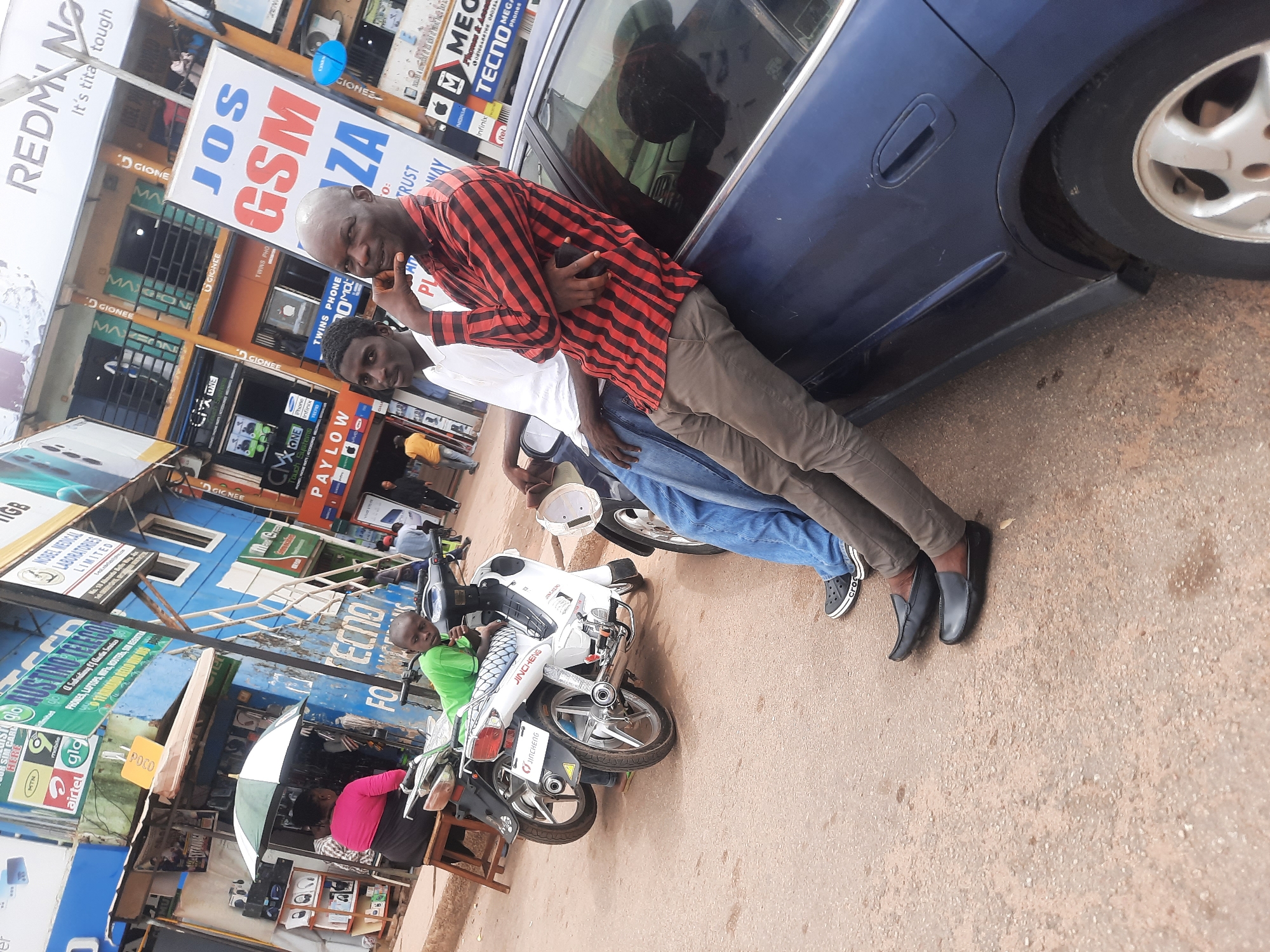 The image shows two men posing in front of a blue car on a street in Jos, Nigeria. The first man, bald and wearing a red and black striped shirt and brown pants, leans against the car. The second man, wearing a white shirt and blue jeans, stands next to him. Numerous storefronts are visible in the background, displaying advertisements for mobile phone brands like Redmi, Gionee, TECNO, and Infinix. Also visible are signs for businesses like JOS GSM ZA and Nobel Medical Laboratories Limited. In the foreground, a man in a green shirt sits on a motorcycle. Another woman sits under an umbrella at a small electronics stand. The street is paved, with visible cracks and unevenness.
