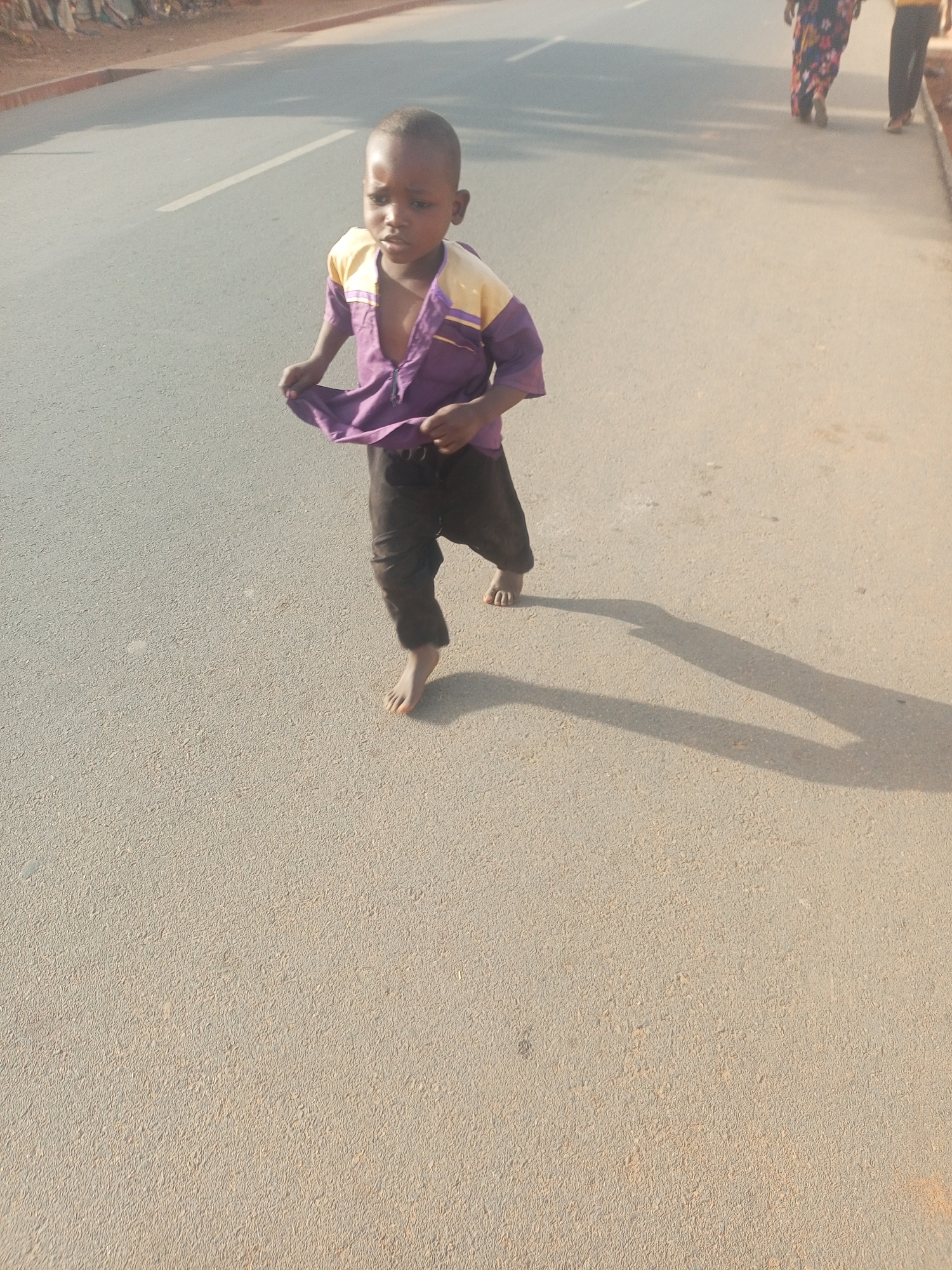 A young, barefoot African boy is walking on a paved road, his feet casting a distinct shadow. He wears a purple shirt with gold accents and dark pants. The road appears to be in a city, though the specific location remains unknown. In the background, two individuals are seen walking away from the camera on the side of the road. The lighting suggests it is daytime. The overall setting seems simple and potentially impoverished.