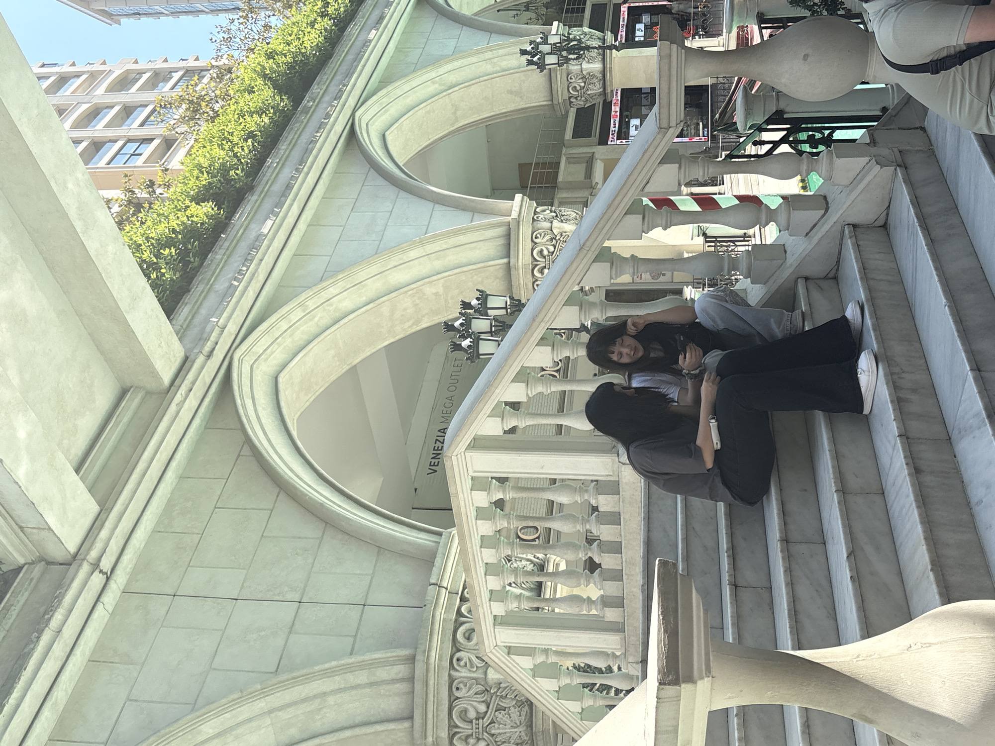 The image depicts a detailed scene at the Venezia Mega Outlet in Gaziosmanpaşa, Turkey, showcasing Venetian-inspired architecture under natural daylight. A grand staircase, constructed from light-colored marble with distinct veining, occupies the central and foreground space. The staircase features elaborate white balustrades with decorative posts and balusters.
Two women are seated on the marble steps. The woman on the left wears a dark grey short-sleeved top, dark-colored flared trousers, and white sneakers. She is looking down, engaged with an object held in her hands. To her right, another woman is seated, wearing a light-colored t-shirt, light blue jeans, and white sneakers. She is actively using a mobile phone, holding it in both hands while resting her chin on her left hand. A third individual, partially visible at the bottom right, wears a light tan t-shirt and has a dark strap across their shoulder.
The background features an expansive building facade characterized by multiple large, rounded arches and decorative columns, constructed from light-colored, block-patterned material. Ornate capitals adorn the columns, and two black, multi-lamp streetlights are mounted on the arch supports. Above the arches, a strip of vibrant green hedges suggests a rooftop garden. Beyond this greenery, modern, multi-story buildings with numerous windows are visible against a clear blue sky. A white sign with black lettering, partially visible behind the arches, reads "VENEZIA MEGA OUTLET." Subtle structural details, such as a thin crack in the stonework above one of the arches, are discernible. Below the stairs and balustrade on the right, a red and white striped pole, reminiscent of a Venetian mooring post, stands next to a glimpse of a blue-green canal or waterway.