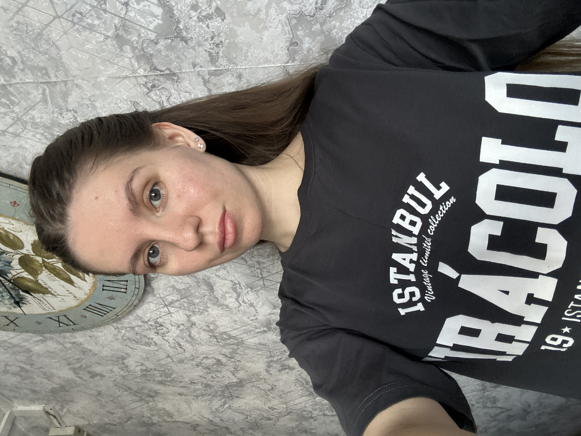A young woman with long brown hair poses for a selfie. She wears a black t-shirt with white print saying "ISTANBUL Vintage limited collection RACCOLI 19*ISTAN". She has fair skin and a small earring in her ear. In the background, a decorative wall clock with Roman numerals is partially visible, and the wall has a textured gray pattern. The location in the city of Klin, Russia is not discernible from the provided image.