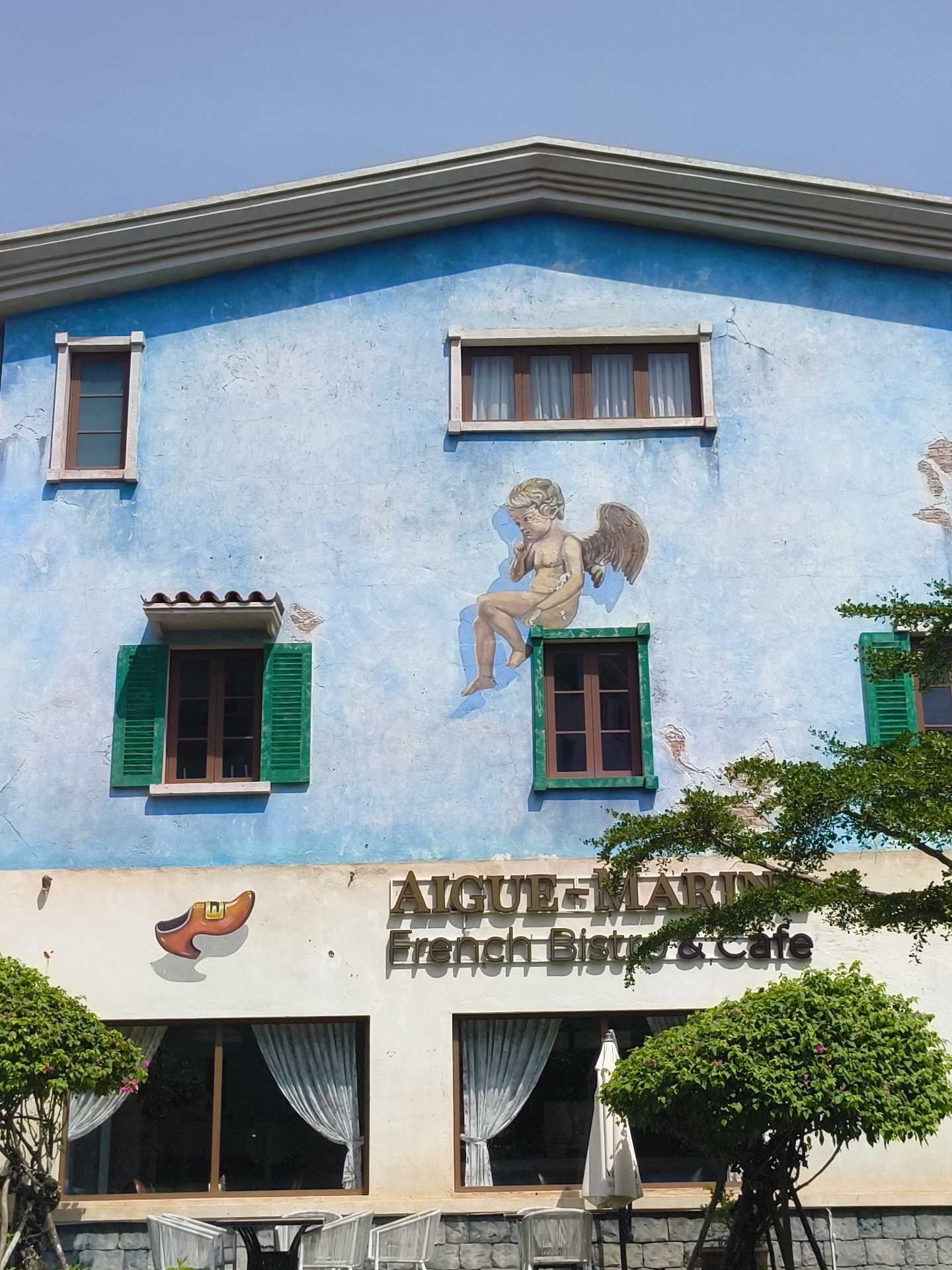 The image shows the exterior of a two-story building with a light blue facade, likely a restaurant or cafe, in Phú Quốc, Vietnam. The building has a pitched roof with white trim. The upper story features a mural of a cherub sitting on a window ledge, and two windows with dark brown frames. One window has green shutters. The lower story has a white facade with large windows covered by white, sheer curtains. Above the windows, a sign reads "AIGUE + MARIN French Bistro & Cafe," with a painting of a wooden shoe to the left of the sign. The building is surrounded by greenery, including a tree with dense foliage and a smaller bush with pink flowers. Outdoor seating with white chairs and tables is visible in front of the building, along with a white umbrella. The sky is clear and blue, indicating a sunny day.